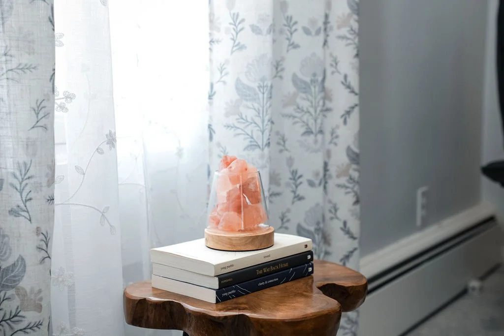 A salt lamp on top of a small stack of books on a wooden side table next to a window with sheer curtains with a floral pattern