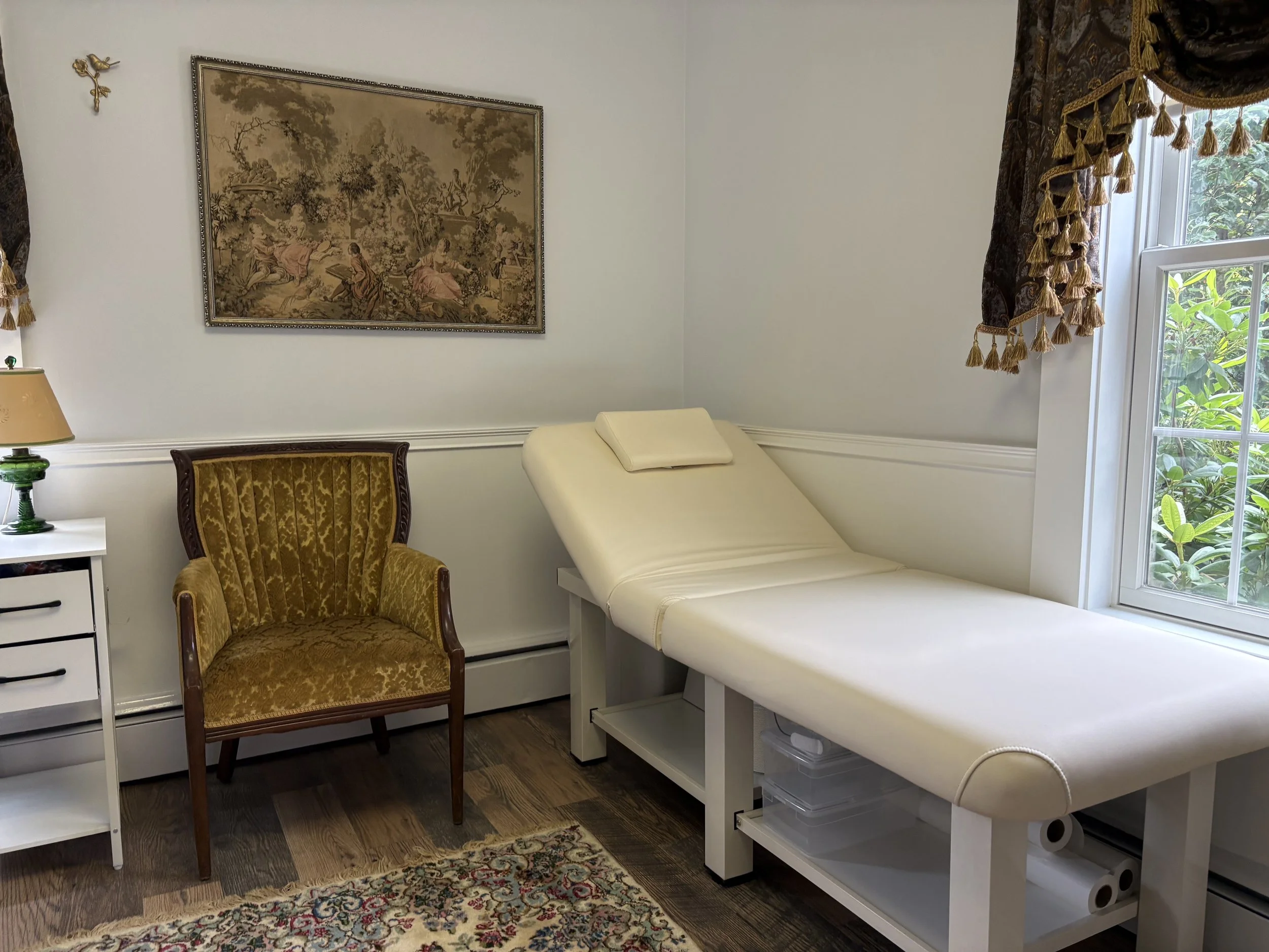 A medical examination room with a padded examination table near a window with ornate dark curtains. There is a vintage-style armchair and a small white side table with a lamp. A large picture with a pastoral scene hangs on the white wall.