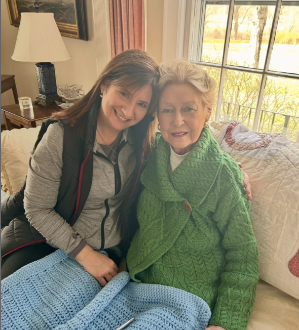 Two women sitting together on a couch, smiling, with a window showing trees and a quilted blanket in the background.