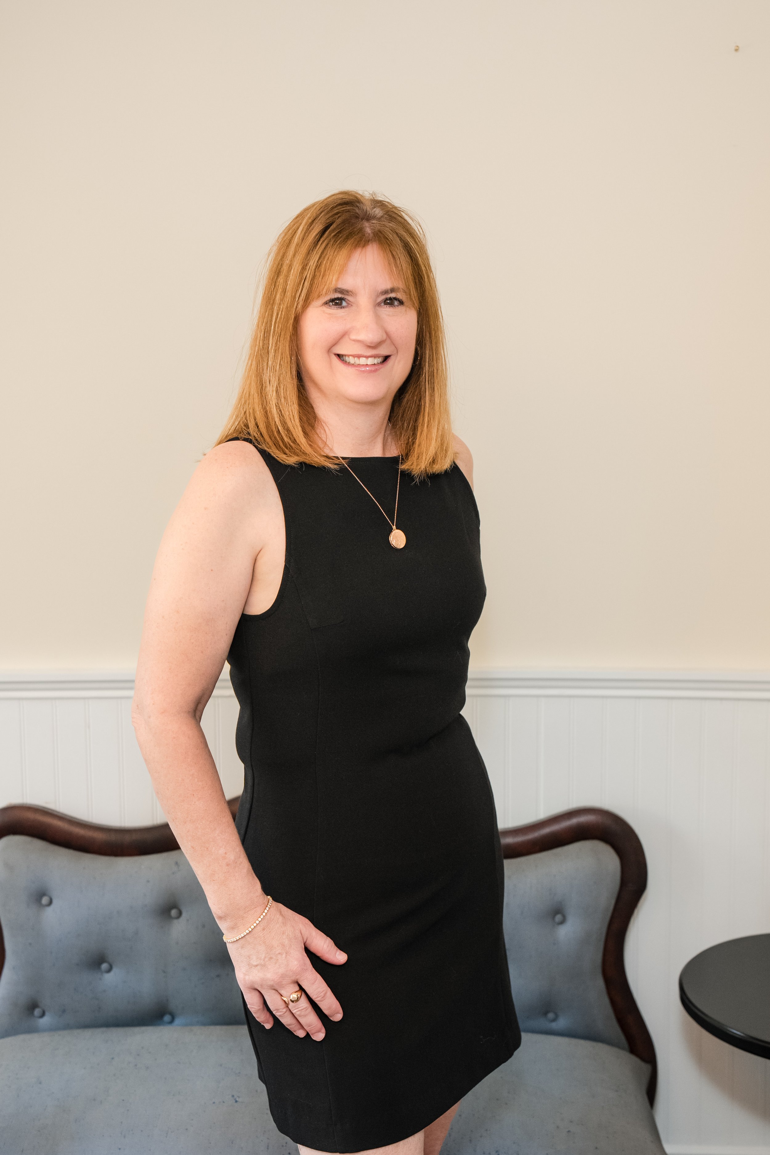 A woman with shoulder-length red hair, wearing a black sleeveless dress and gold jewelry, smiling and standing in front of a beige wall and a gray upholstered chair with dark wood framing.