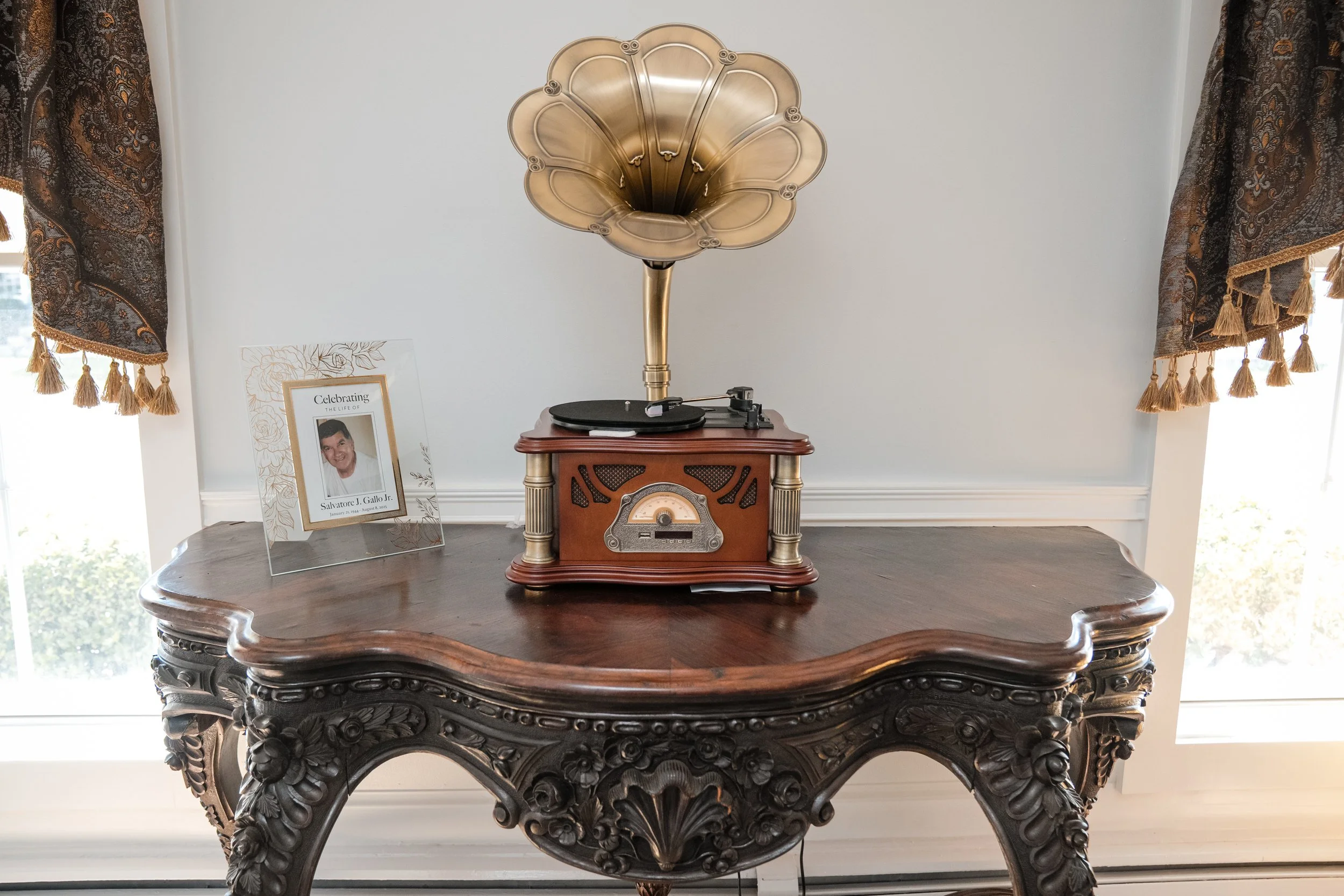 Vintage music player with a carved wooden base and a brass horn, placed on an ornate wooden table, with floral curtains on either side and a dividend picture frame on the left.