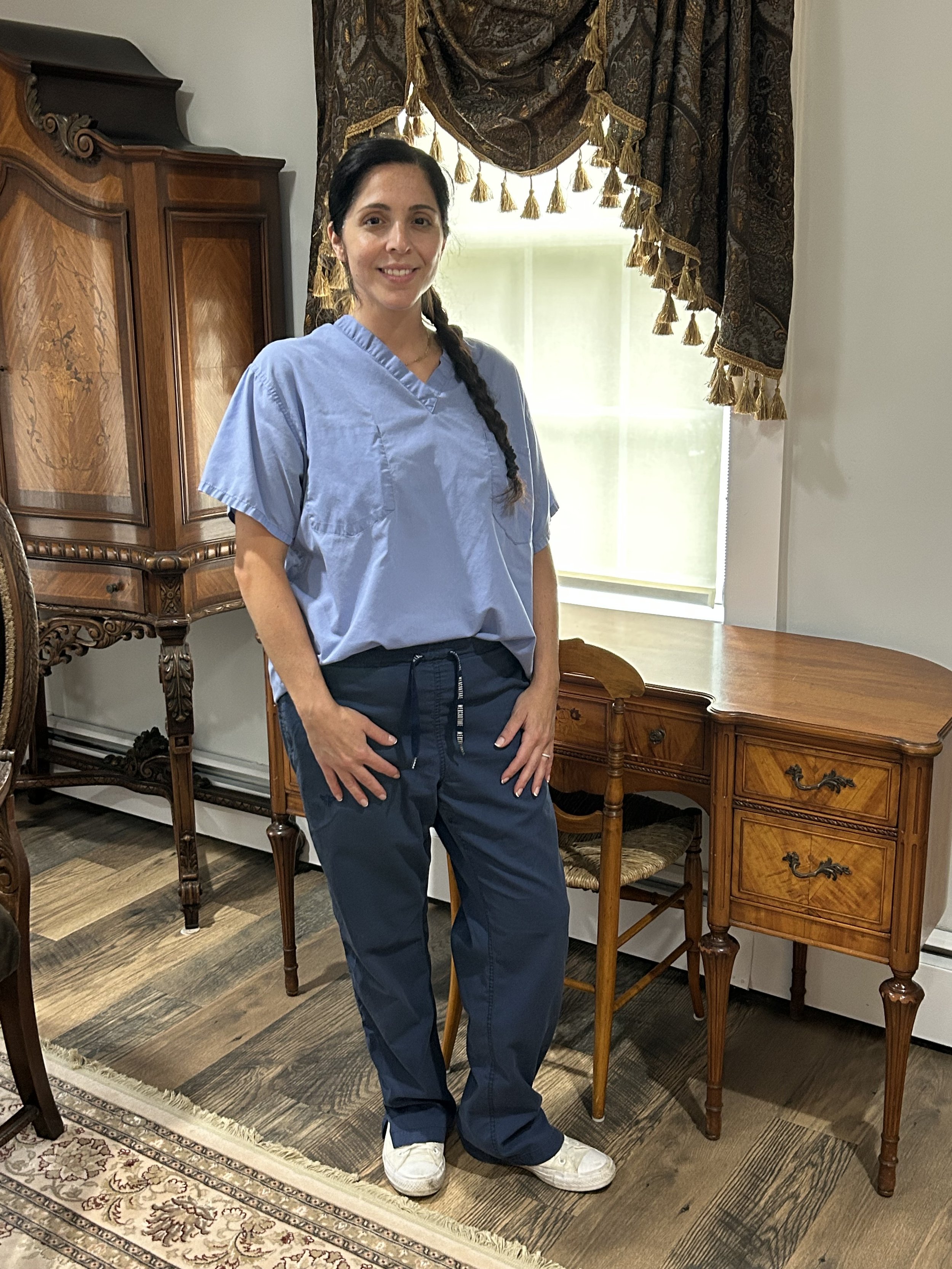 A young woman with dark hair in a braid, wearing a light blue scrub top, navy pants, and white sneakers, standing in a room with wooden furniture, a large window with drapes, and a patterned rug.