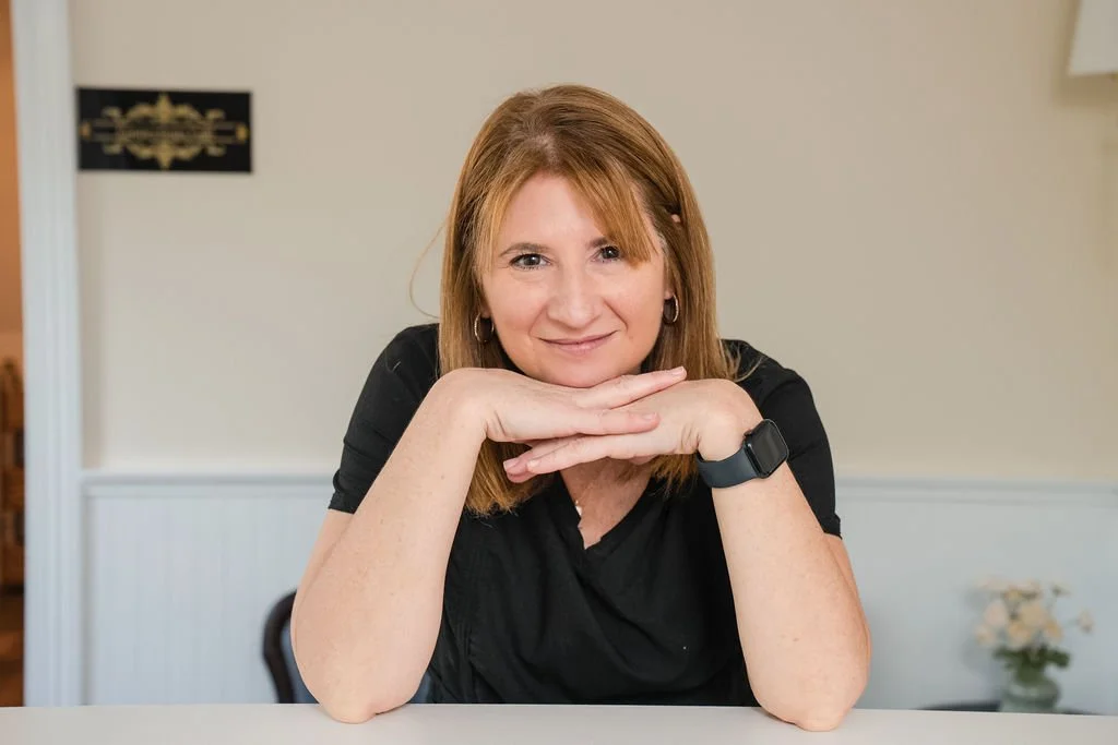 A woman with shoulder-length reddish hair wearing a black top and a smartwatch, sitting at a table with her chin resting on her hands, smiling at the camera.