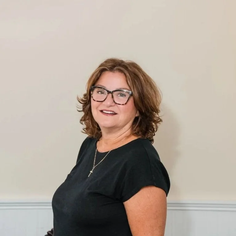 A woman with shoulder-length curly brown hair and glasses is smiling at the camera, wearing a black shirt and a gold cross necklace, standing against a plain light-colored wall.