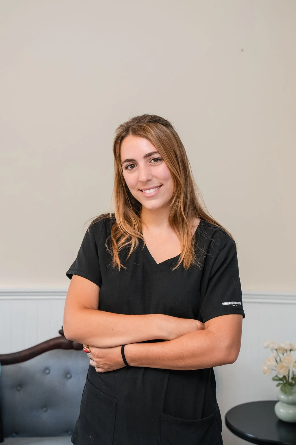 A woman with light brown hair wearing black scrubs, smiling, arms crossed, standing in a room with a light-colored wall, a blue upholstered bench, and a small table with a vase of white flowers.