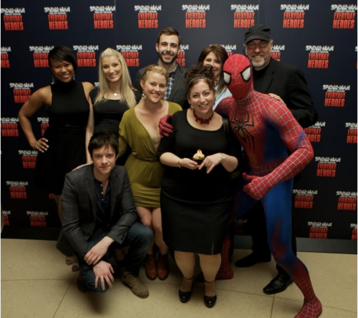 A group of nine people, including a person dressed as Spider-Man, posing in front of a backdrop that features the 'Spider-Man: Into the Spider-Verse' and 'Everyday Heroes' logos.
