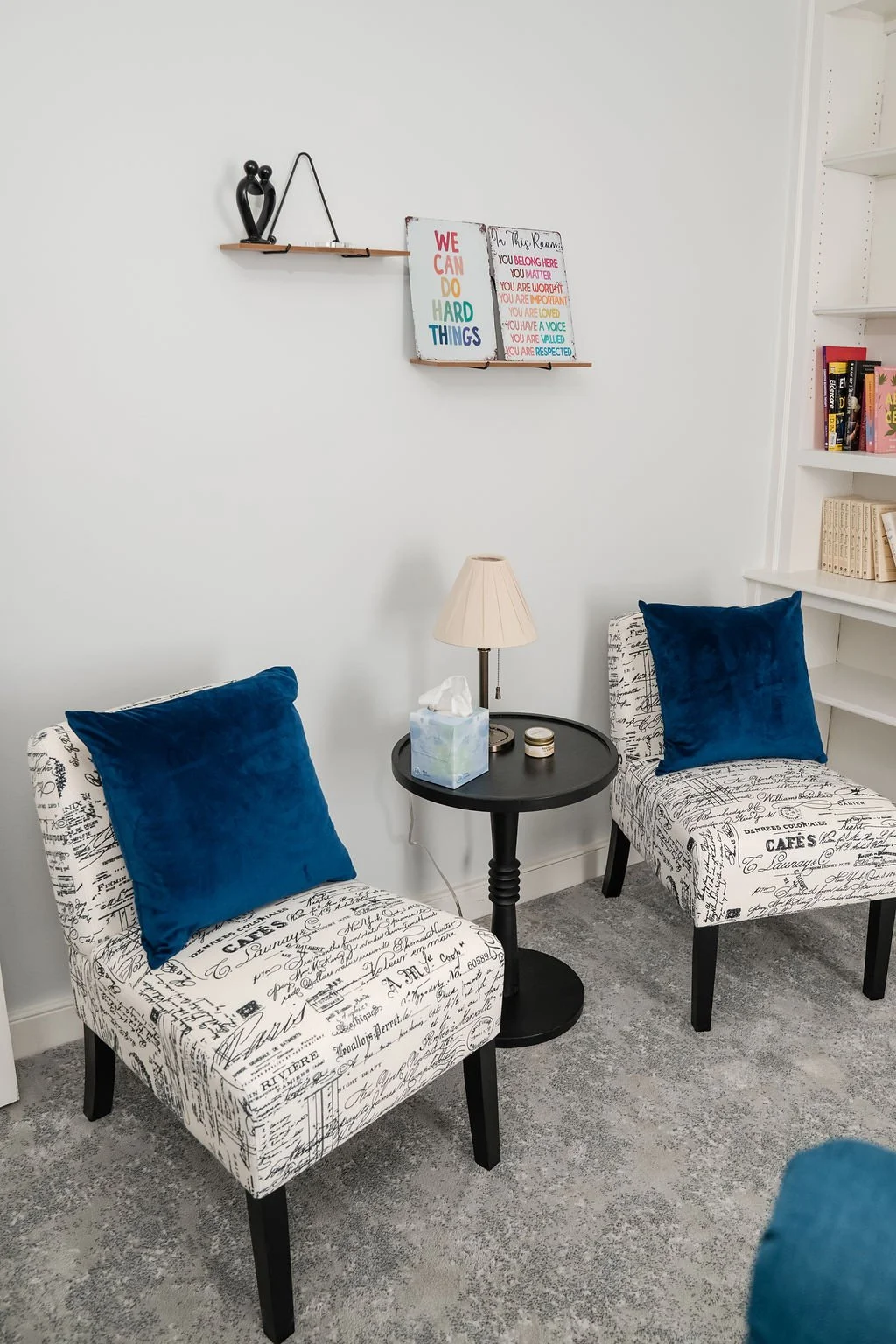 A cozy seating area with two patterned chairs with black writing scripts, each adorned with a royal blue velvet pillow, positioned on either side of a small black round side table with a beige lamp on top. Above, there are two shelves with decorative items and framed posters on a white wall; the posters contain colorful motivational quotes. To the right, part of a white bookshelf with books is visible.