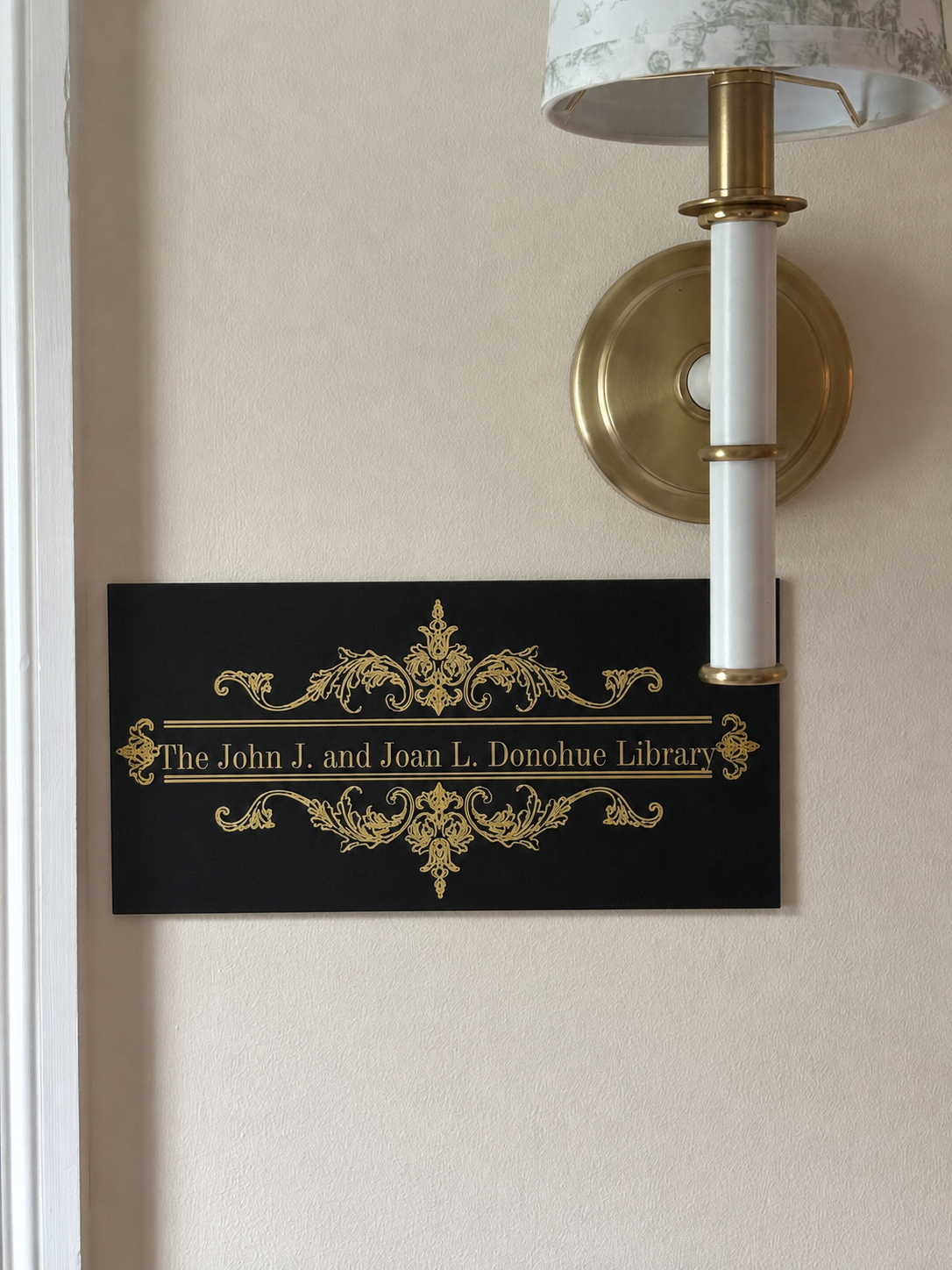 A black sign with gold decorative details and text that reads "The John J. and Joan L. Donohue Library" mounted on a beige wall. A brass wall sconce with a light-colored lampshade is partially visible above the sign.