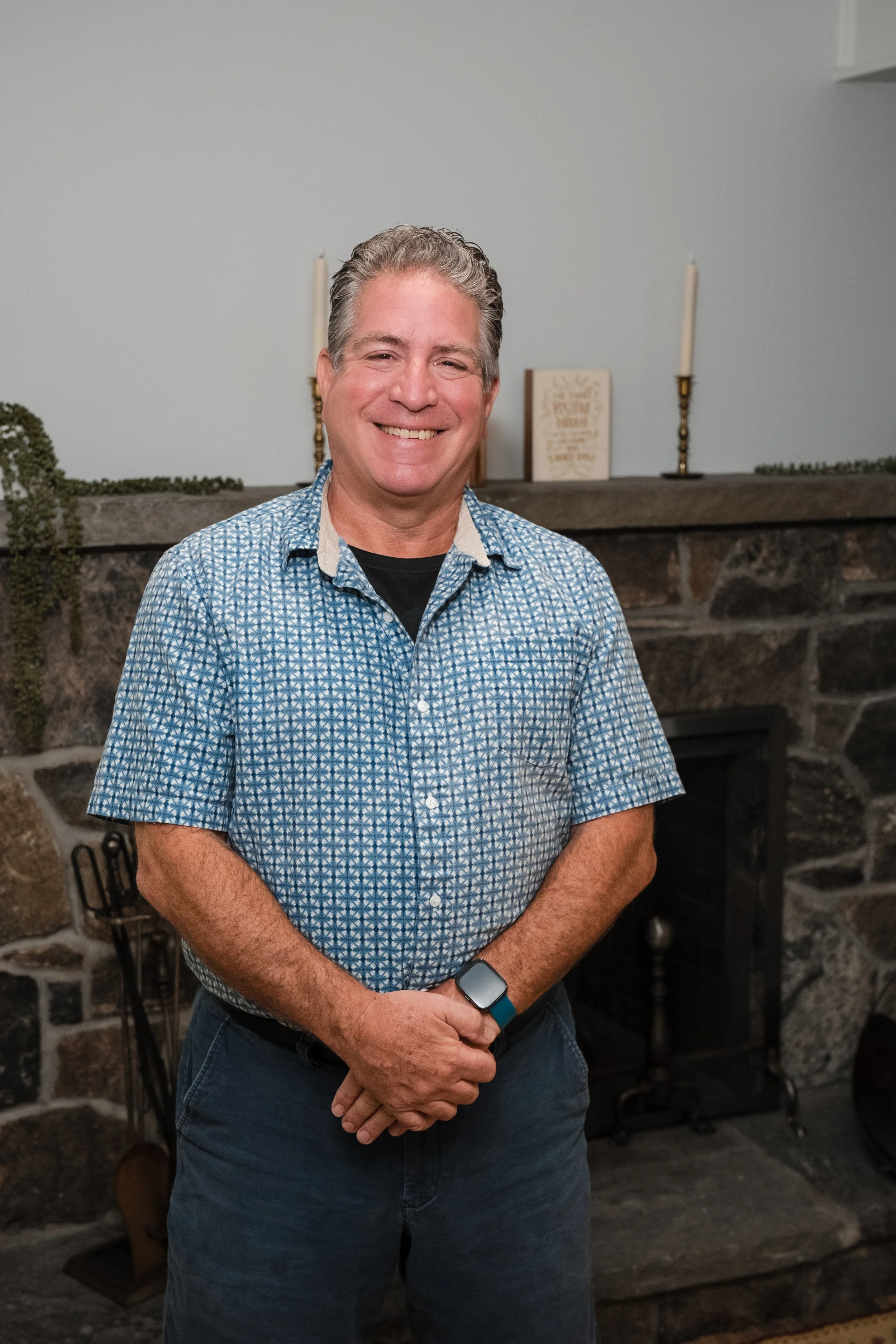 A smiling man with gray hair wearing a patterned blue shirt and a smartwatch, standing in front of a fireplace with candles and decorative books.