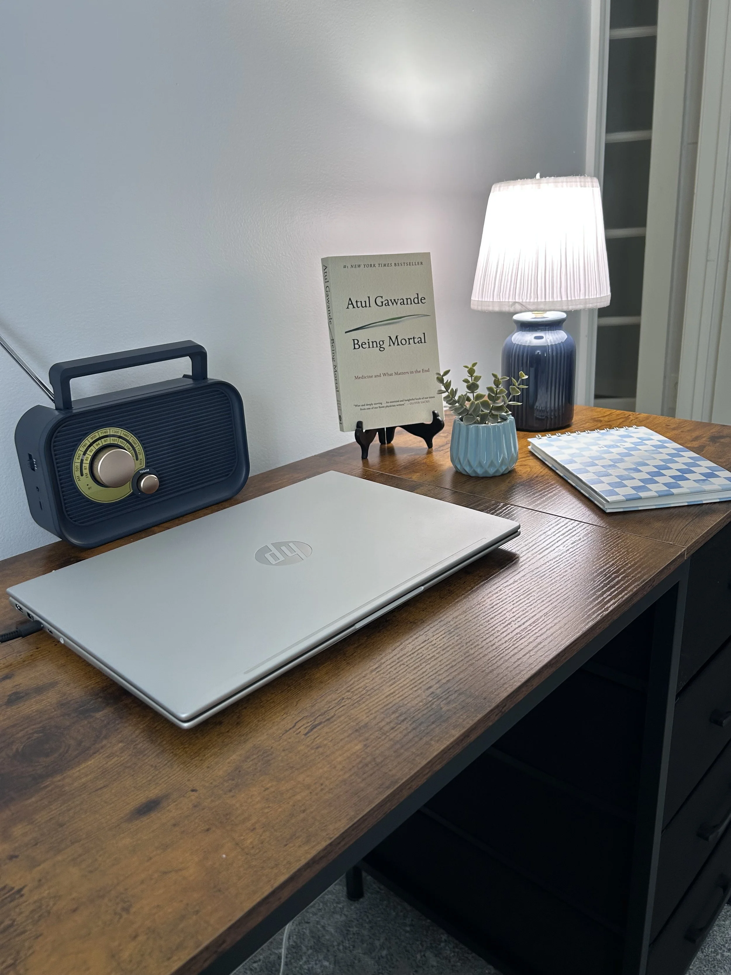 A wooden desk with a closed silver HP laptop, a vintage blue radio, a small potted plant in a blue pot, a white and blue checkerboard notebook, a table lamp with a white shade, a book titled 'Being Mortal' by Atul Gawande on a small wooden stand, and a white wall with a closet door in the background.