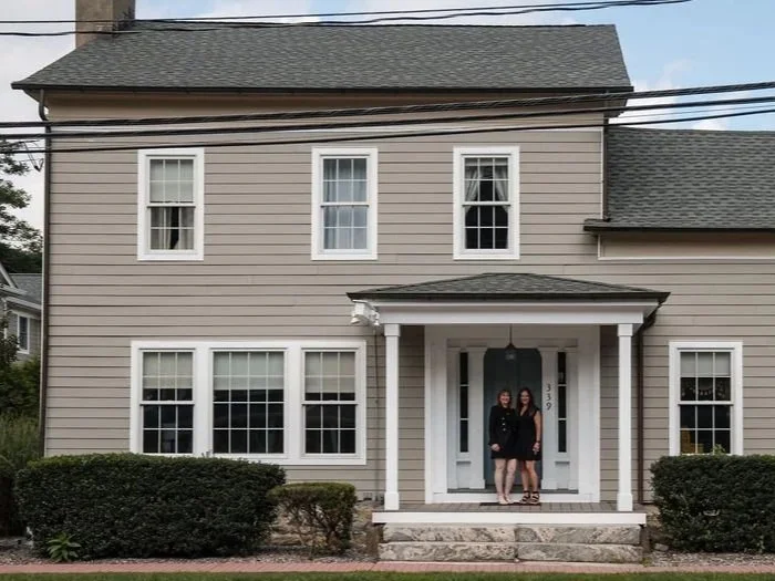 Two women standing in front of a two-story beige house with white trim and a front porch.