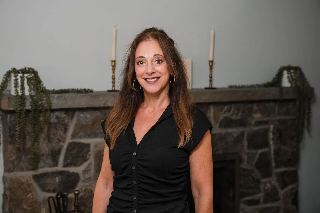 A woman with long brown hair, wearing a black short-sleeved shirt, standing in front of a stone fireplace with a mantle decorated with candles and greenery.