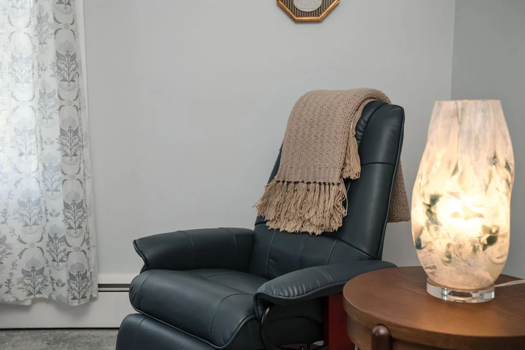 A black leather recliner with a beige knit throw blanket draped over its back, positioned next to a round wooden table with a glowing marble-patterned lamp on it. A white curtain with gray floral patterns is visible on the left side of the image, and a small octagonal wall clock hangs above the recliner.
