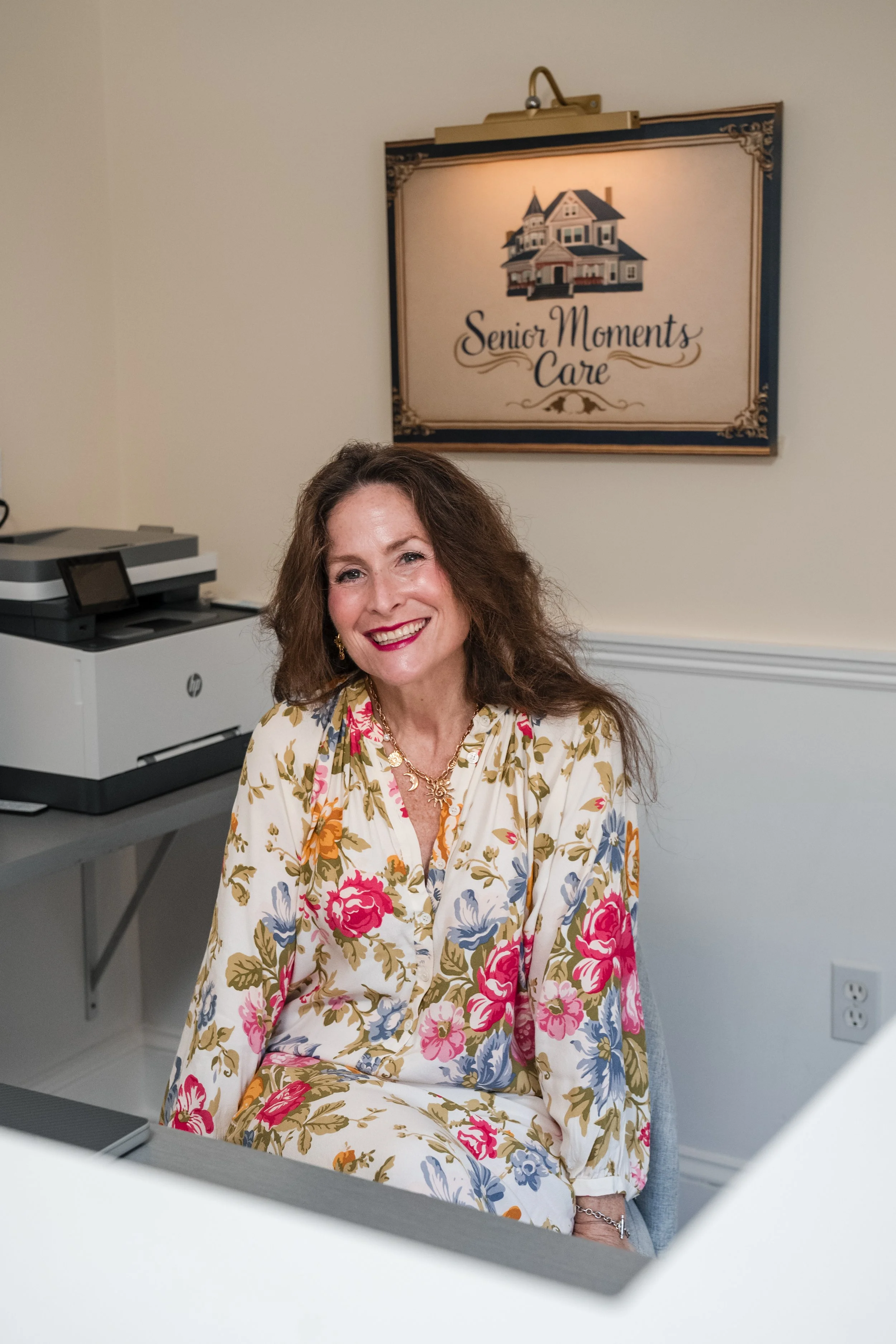 A smiling woman with brown hair, wearing a floral dress, sitting in an office with a sign that reads "Senior Moments Care" and a picture of a house on the wall behind her.