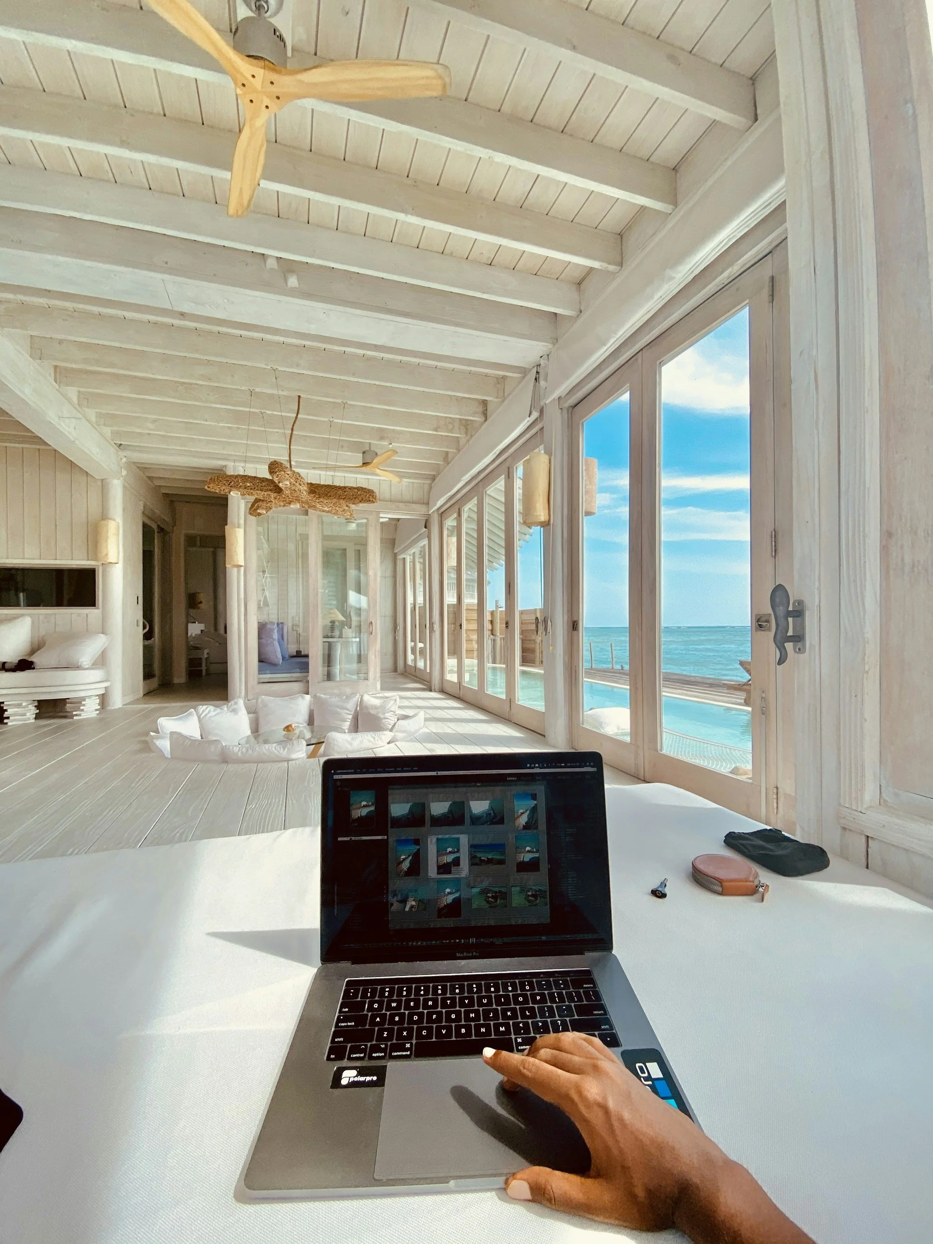 A person using a laptop at a white desk with ocean views through large glass windows, featuring white wooden walls and ceiling, a ceiling fan, and tropical decor.