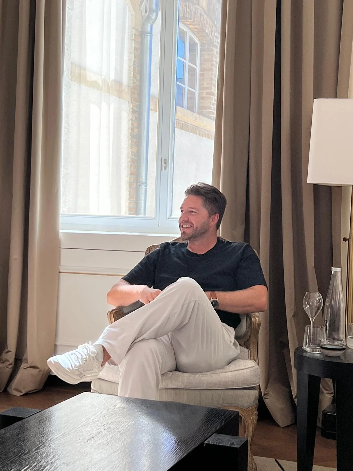 A man sitting on an armchair near a window with curtains, smiling and looking to the side. There is a table with glasses and a water bottle nearby.