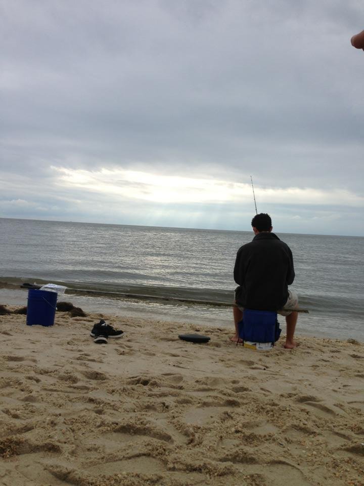 A person sitting on a small bench at the beach, fishing in the ocean on a cloudy day. There are shoes on the sand and a blue container nearby.