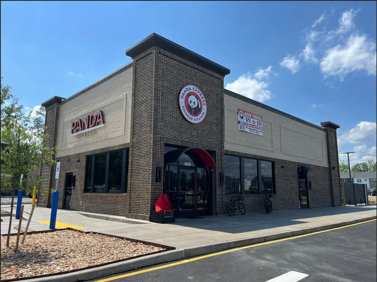The exterior of a Panda Express restaurant with brick walls, large windows, and a circular panda logo above the entrance. A sign indicates the restaurant is hiring, and there are bicycles parked outside.