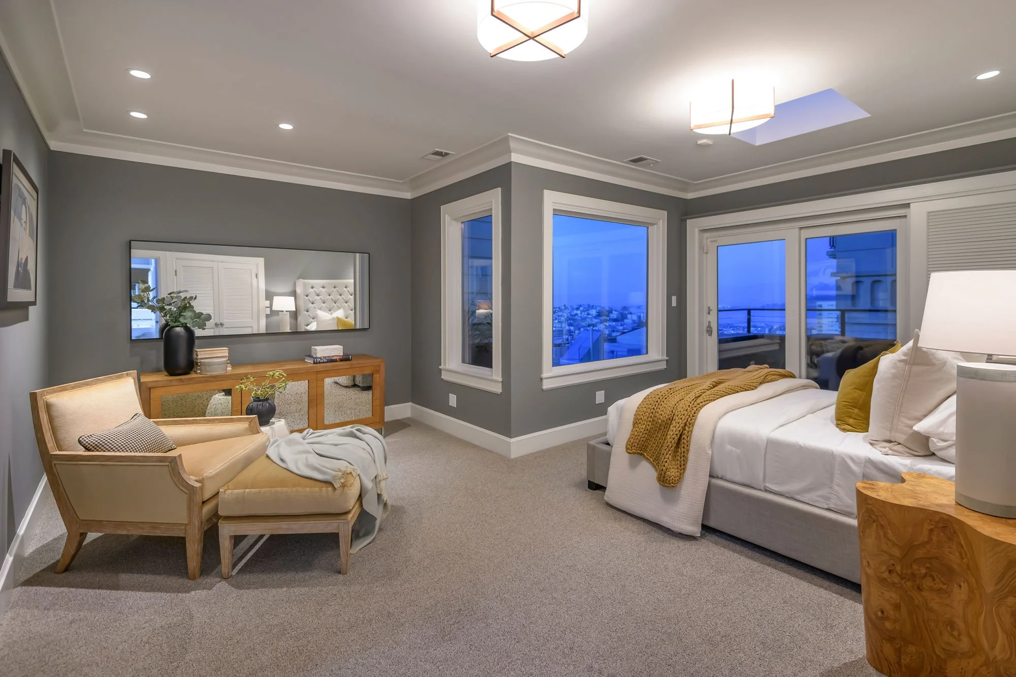 A bedroom with a bed, white bedding, and yellow pillows by a large window showing a city view. There is a wooden nightstand, a beige upholstered armchair with a throw blanket and cushions, a wooden console table with a black vase and decorative items