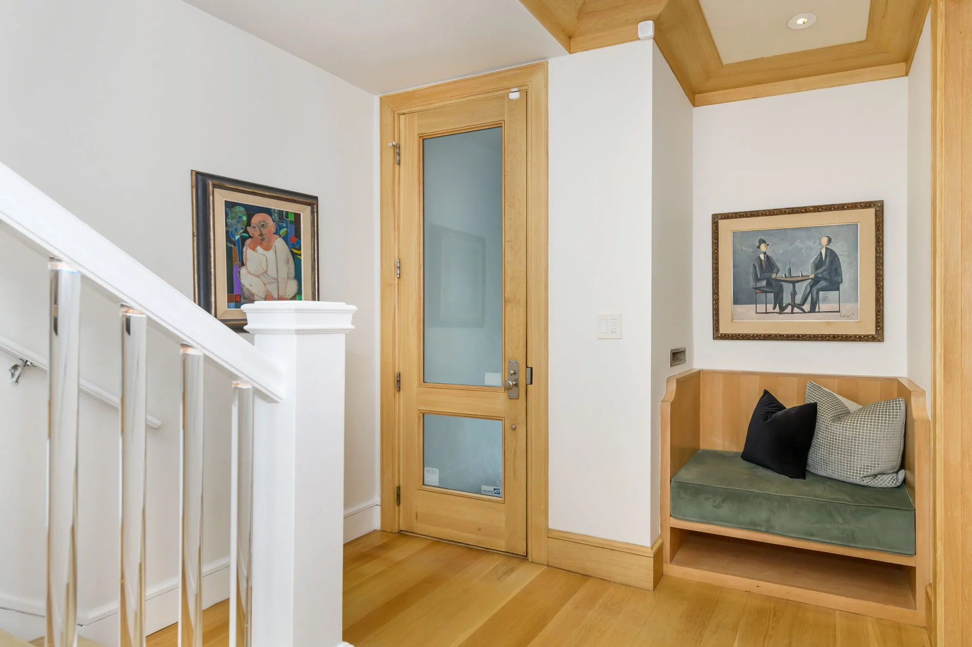 Interior view with a light wooden front door with frosted glass panes, colorful artwork on the wall to the left, a small built-in wooden seating nook with cushions on the right, and white walls and ceiling. Part of a staircase with white railing is v
