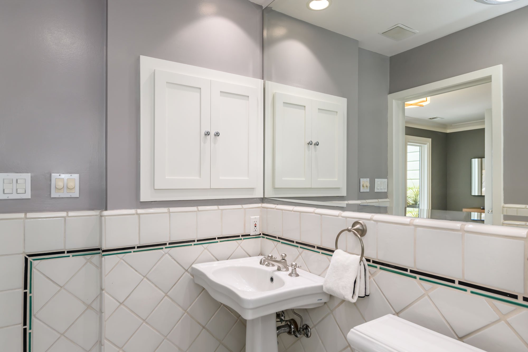 Bathroom with light gray walls, white tile backsplash with decorative black and green border, farmhouse sink, and white cabinets reflected in mirror.