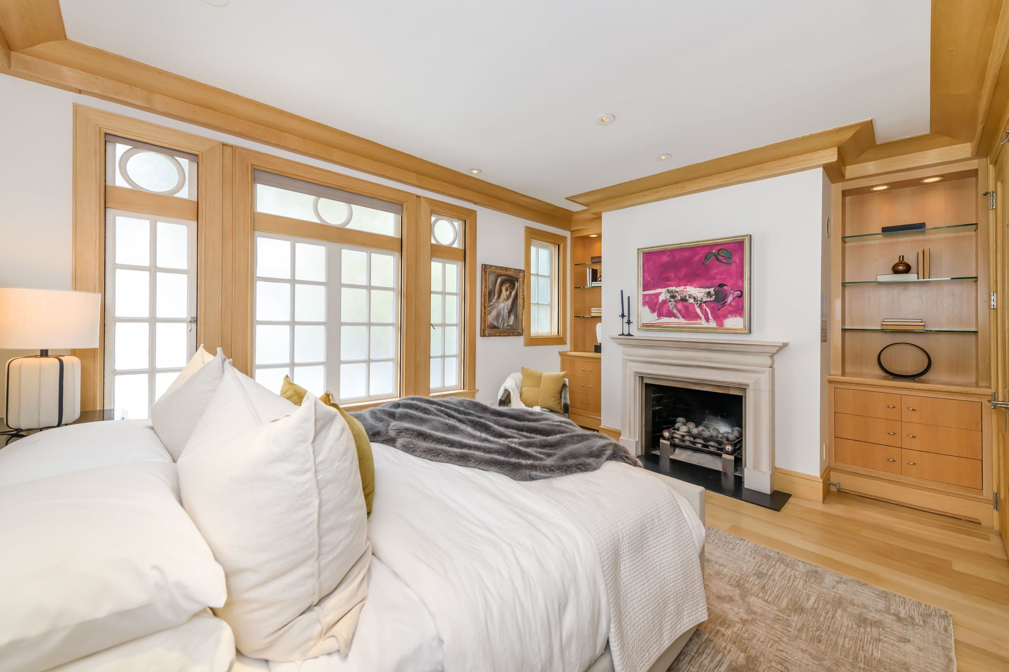 A cozy bedroom with white bedding, a faux fur throw, wooden accents, and artwork on the walls, including a pink abstract painting and a portrait. Large windows with frosted glass let in natural light, and a fireplace with decorative stones is visible
