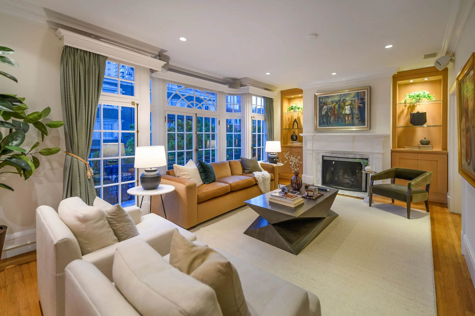 Living room with large windows, green curtains, beige walls, cream and brown furniture, a fireplace, and wall art.