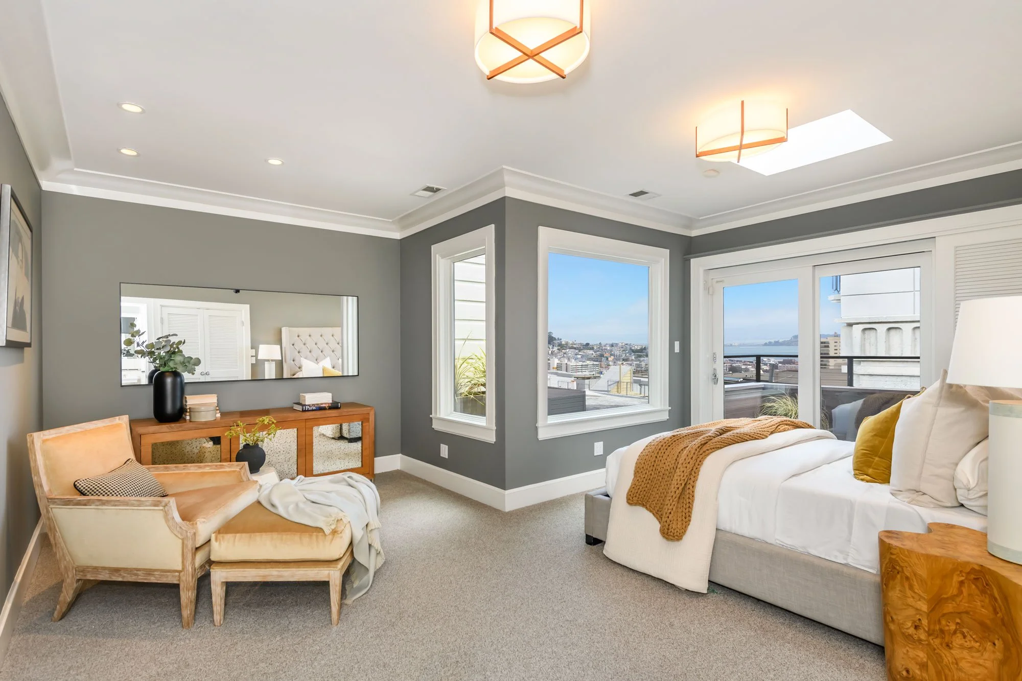 A modern living room with gray walls, large windows showing a city view, a white sofa with yellow pillows, a wooden side table, a vintage armchair, and ceiling lights.