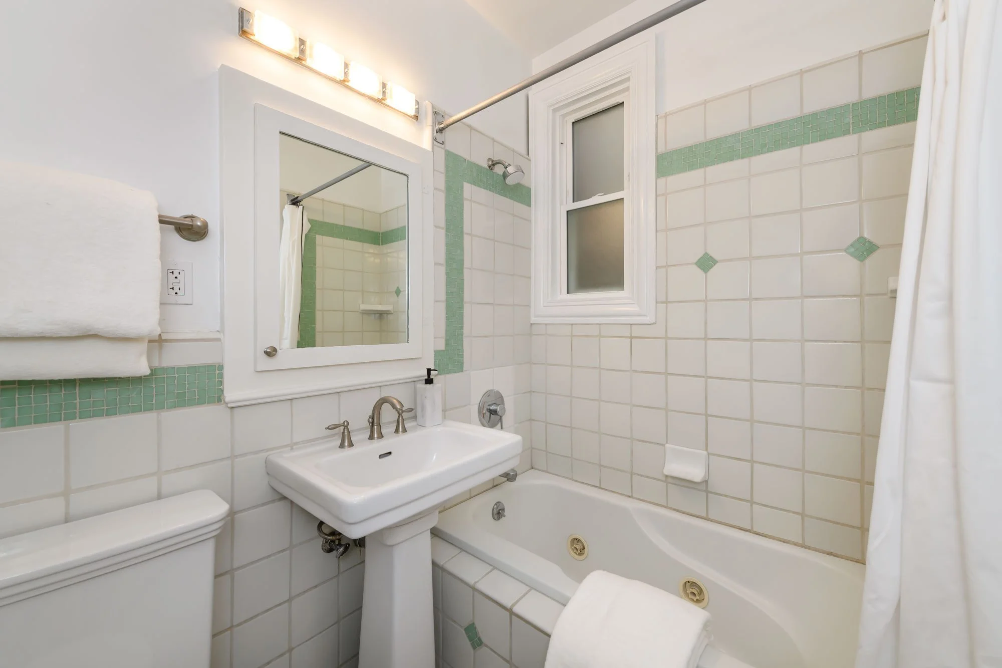 Bathroom with white tiles with green accents, a small window, a bathtub, a toilet, a sink, a mirror, a towel, and a frosted window.