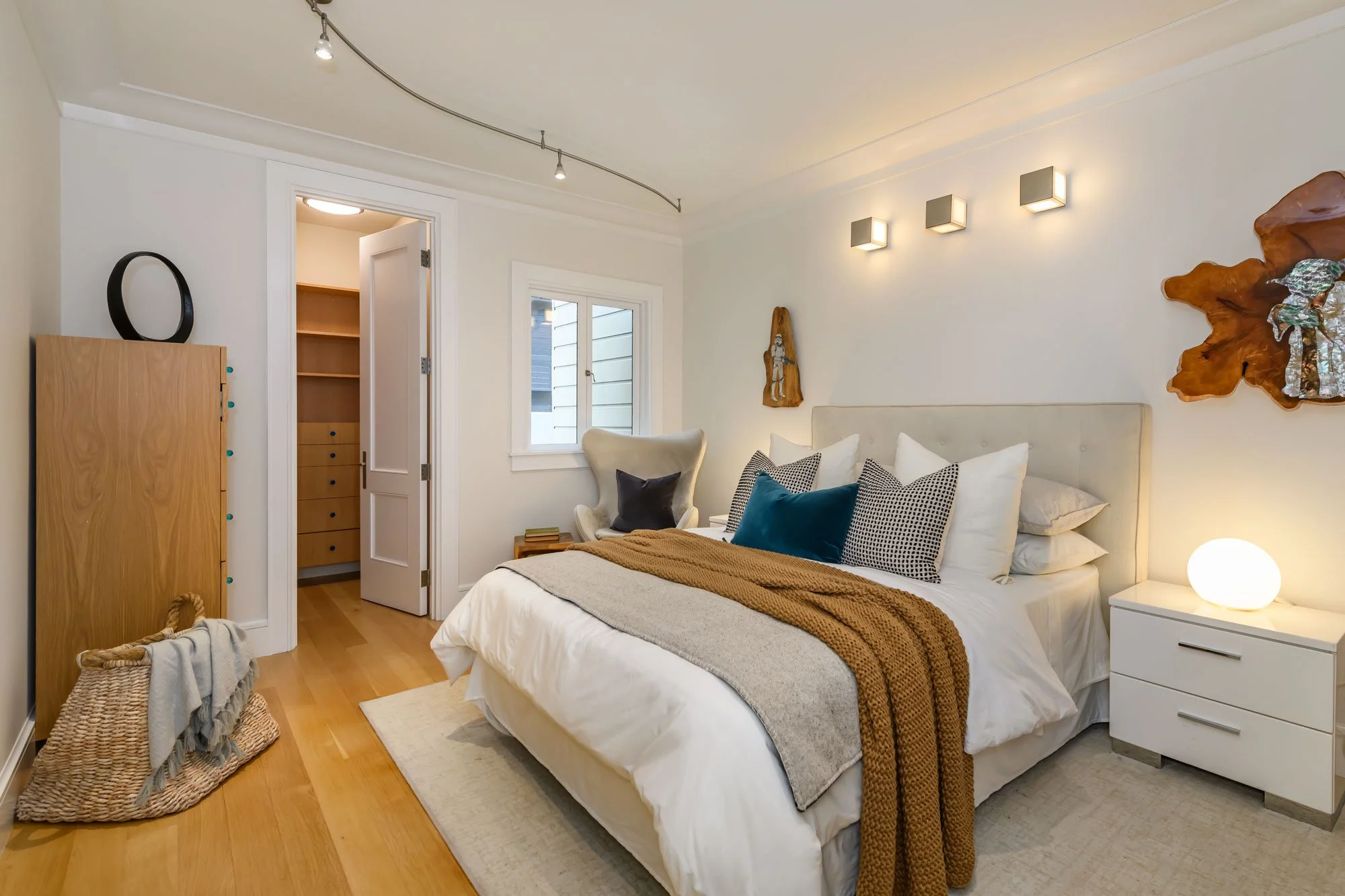 A cozy bedroom with a large bed, pillows, and blankets. There is a nightstand with a spherical lamp, artwork on the wall, a window, and a closet door. The room has wooden flooring and modern lighting fixtures.