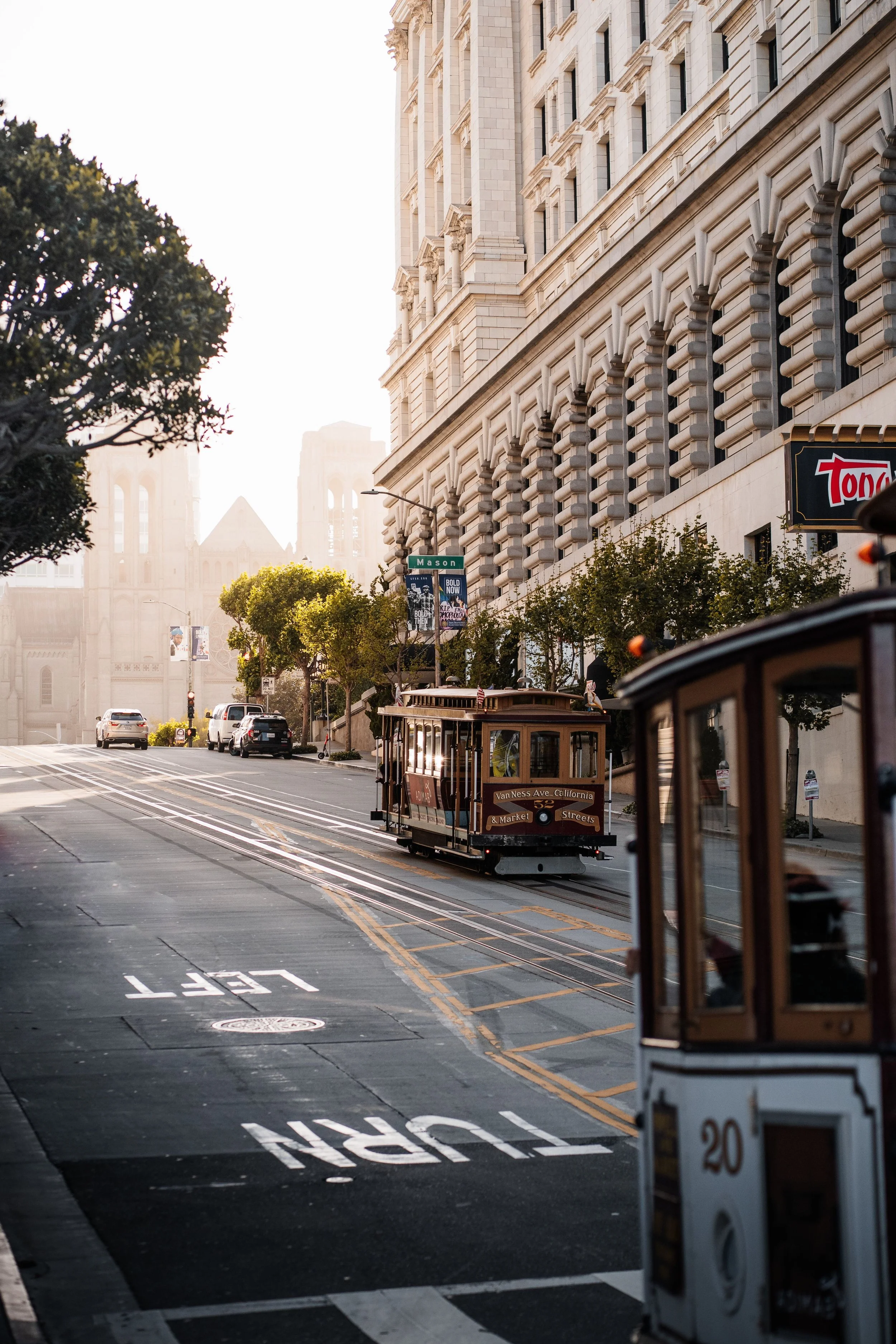 A city street scene with a historic building on the right, a cable car on the tracks, and a church with twin towers in the background. The street has trees, cars, and street signs, with sunlight shining through.