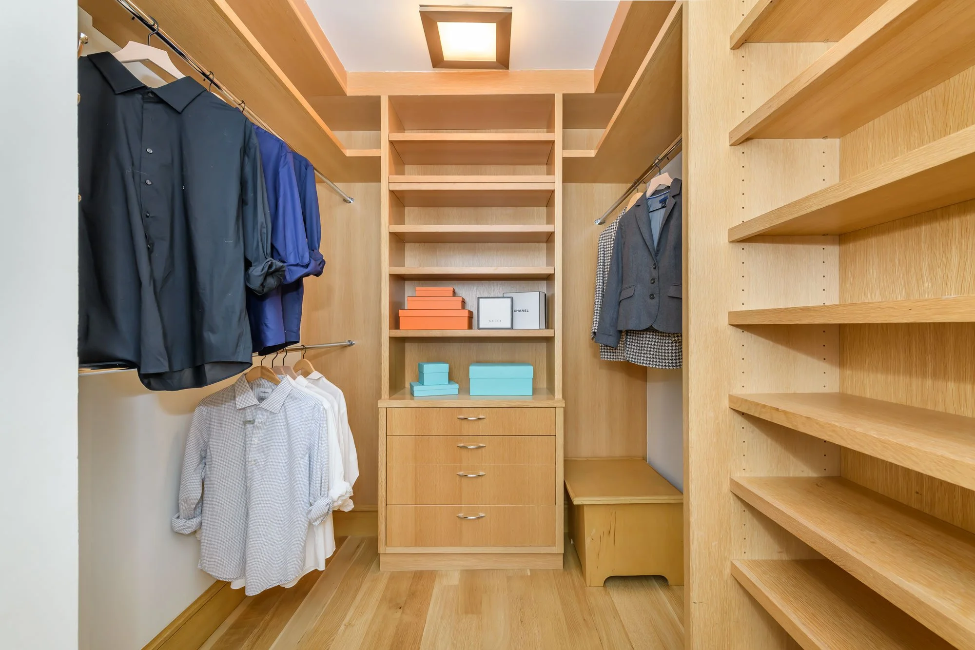 A walk-in closet with wooden built-in shelves and drawers, with hanging clothes on both sides, including black, blue, gray suits, and white shirts. There are two white boxes on the middle shelf, and orange boxes on the lower shelf. Wooden flooring an