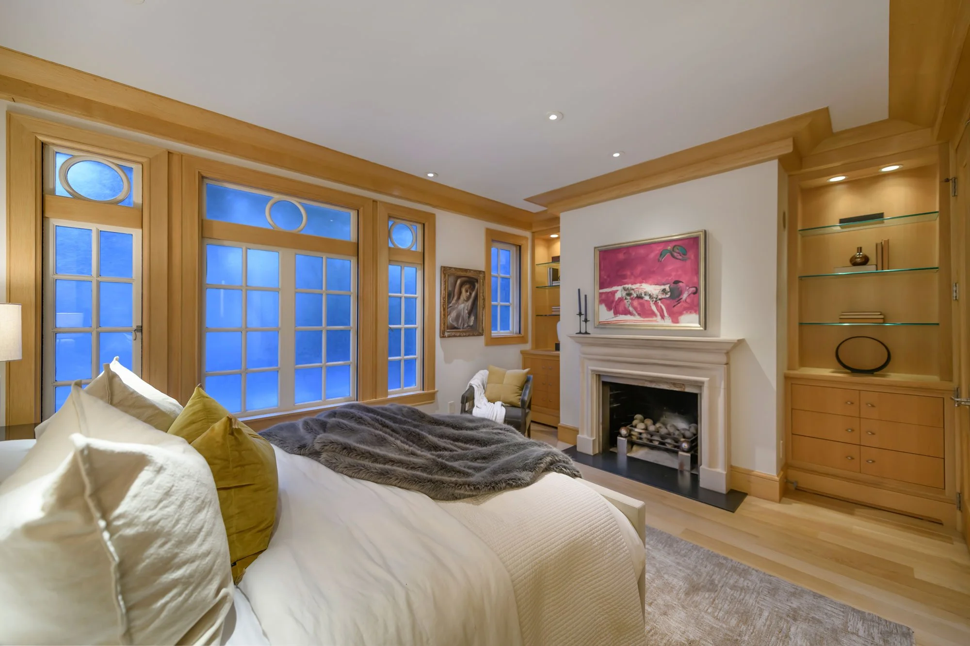 Living room with wooden window frames, white walls, fireplace, artwork, built-in shelves, and a bed with white and yellow pillows and a gray blanket.