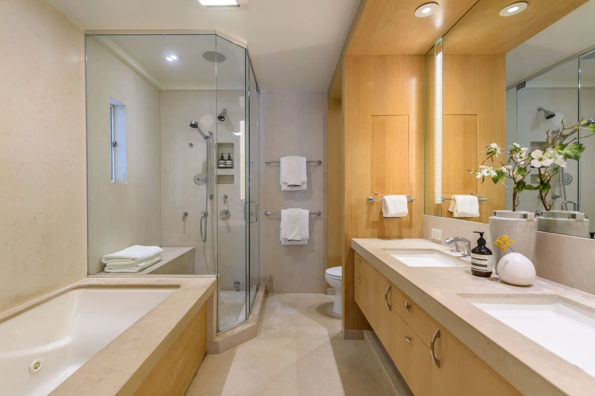 Modern bathroom with a glass-enclosed shower, a built-in bathtub, light wood cabinetry, a large mirror, and decorative plants on the countertop.