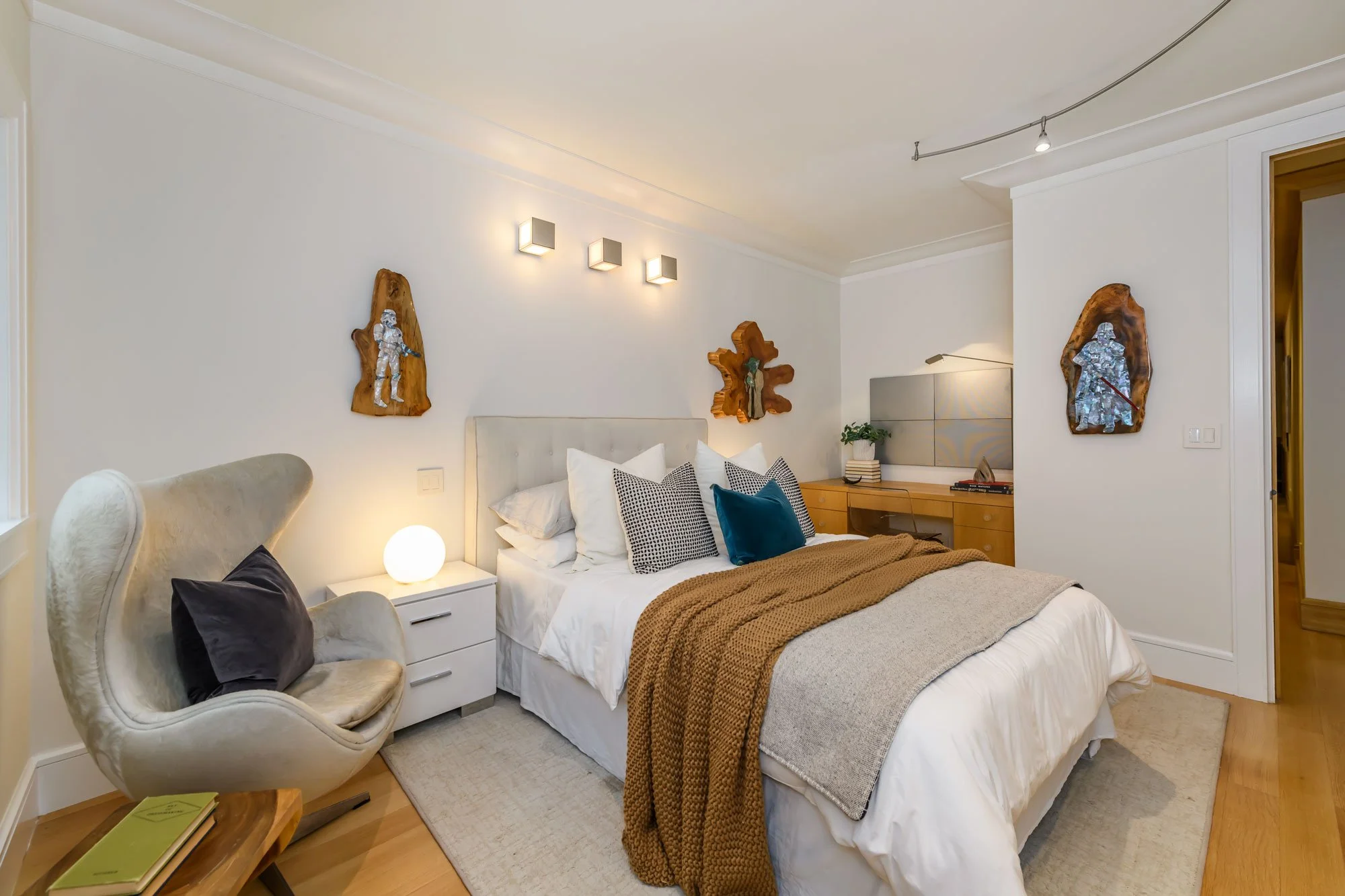 A bedroom with a white bed, decorative pillows, and a brown blanket. Wall art includes wooden plaques with metallic figures. A cream armchair with dark pillows is beside a small wooden table with green books. Modern light fixtures are on the wall, an