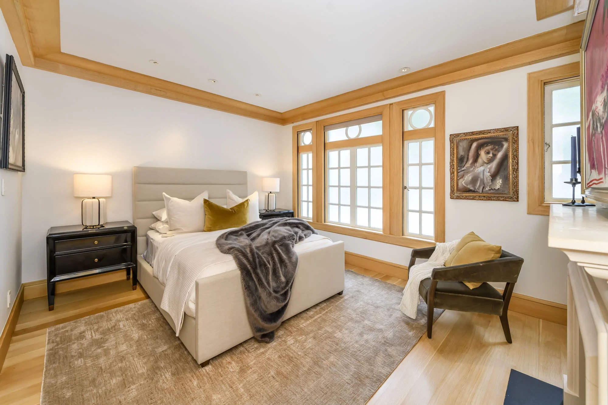 A bedroom with a beige upholstered bed, black nightstands with lamps, large windows with wooden framing, artwork on the walls, a beige rug, and a wooden floor.