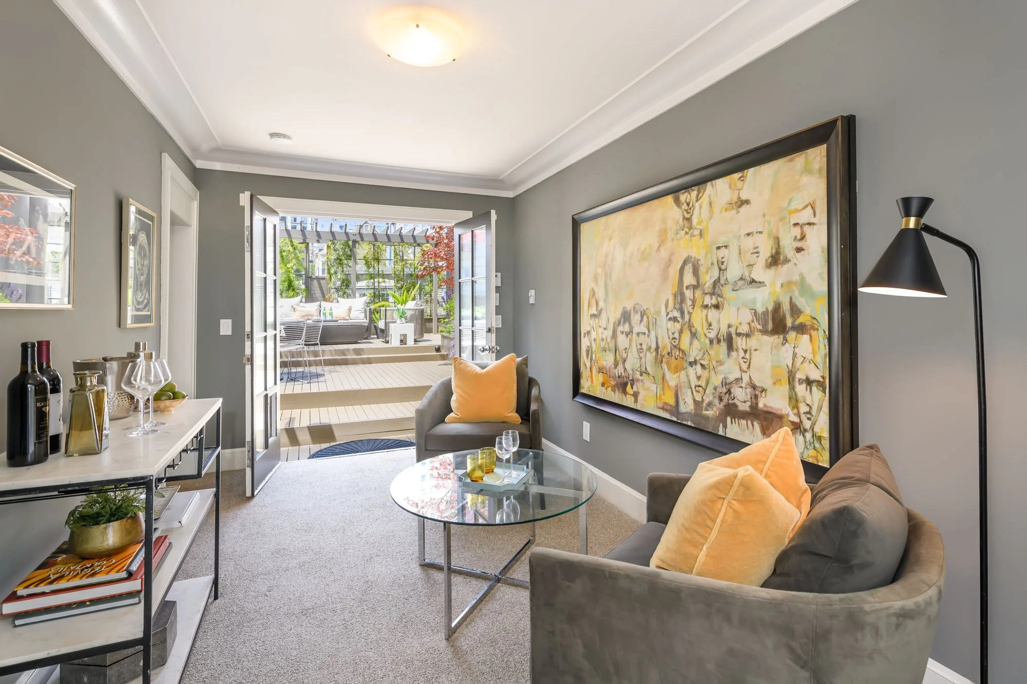 Living room with gray walls, large abstract painting, glass coffee table, beige and yellow cushions, floor lamp, and a door leading to a patio with outdoor seating and greenery.