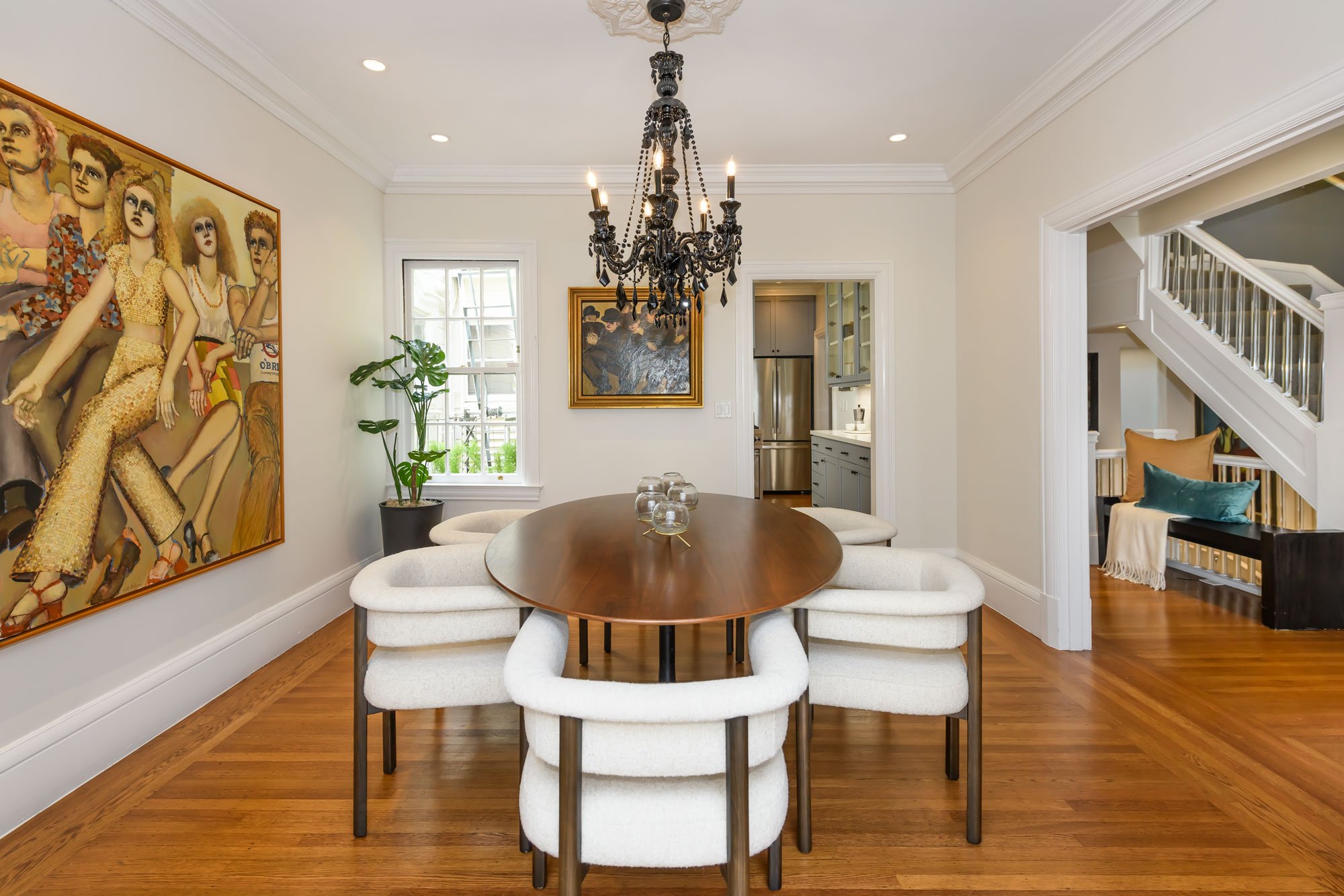 A dining room with a round wooden table surrounded by six white upholstered chairs. A black ornate chandelier hangs above the table. There is artwork on the walls, and a window with a potted plant nearby. The background shows a kitchen and a staircas