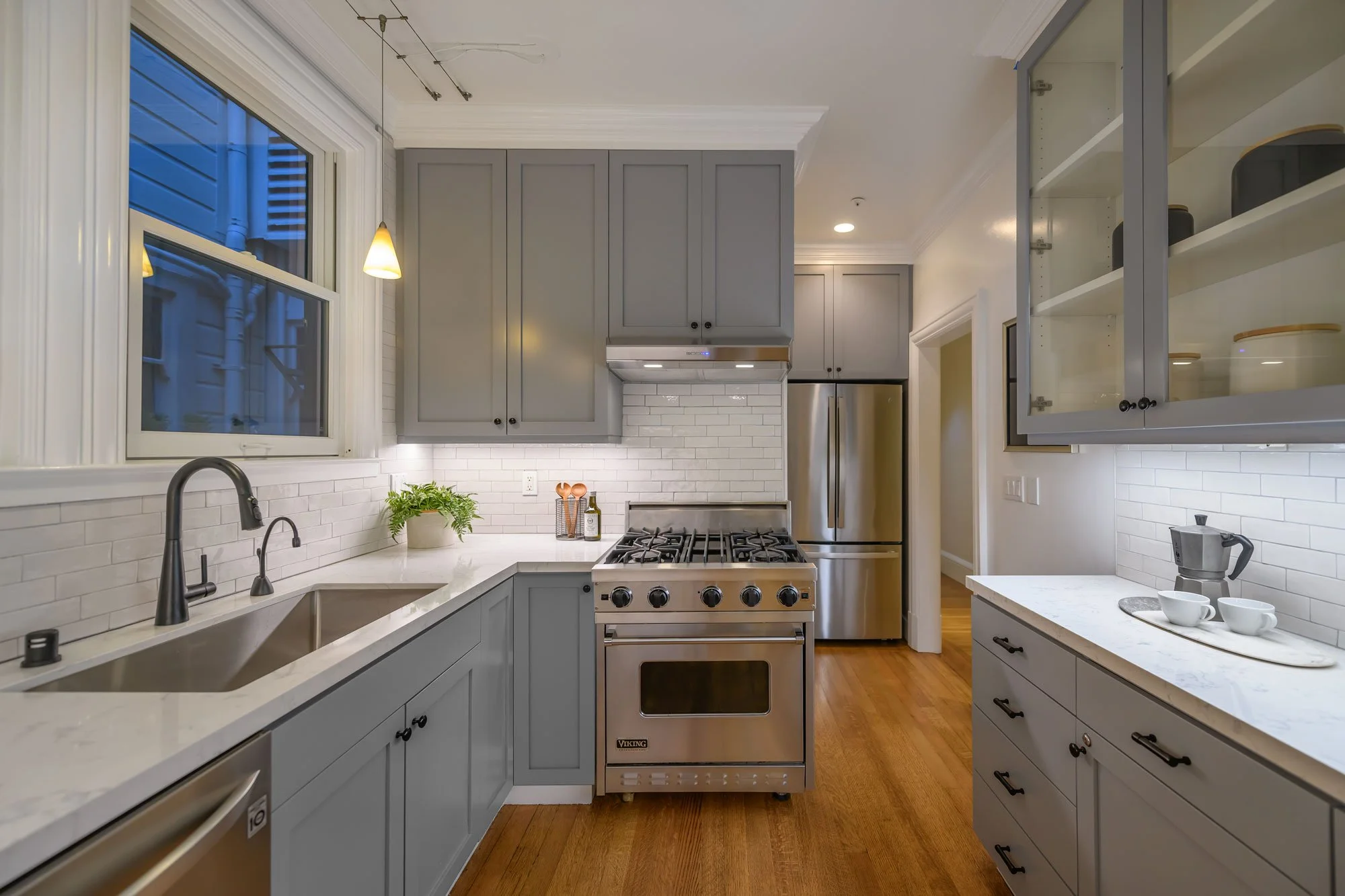 Modern kitchen with light gray cabinets, white subway tile backsplash, stainless steel appliances including a gas stove and refrigerator, white countertops, a window above the sink, and small hanging pendant lights. There are some potted plants and k