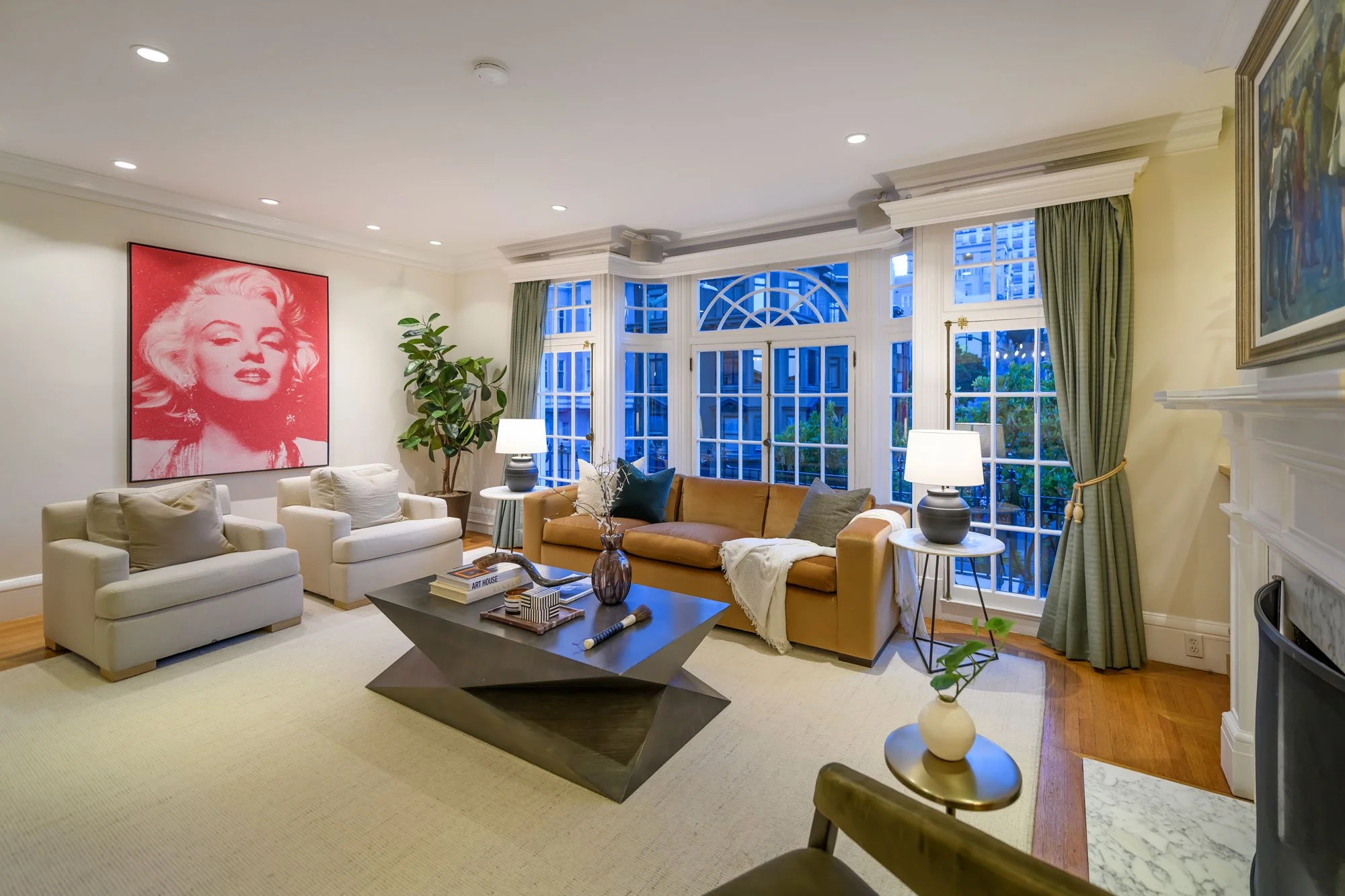 Living room with white sofas, a mustard couch, a black coffee table, art on the walls, large bay windows with green curtains, lamps, plants, and decorative items.