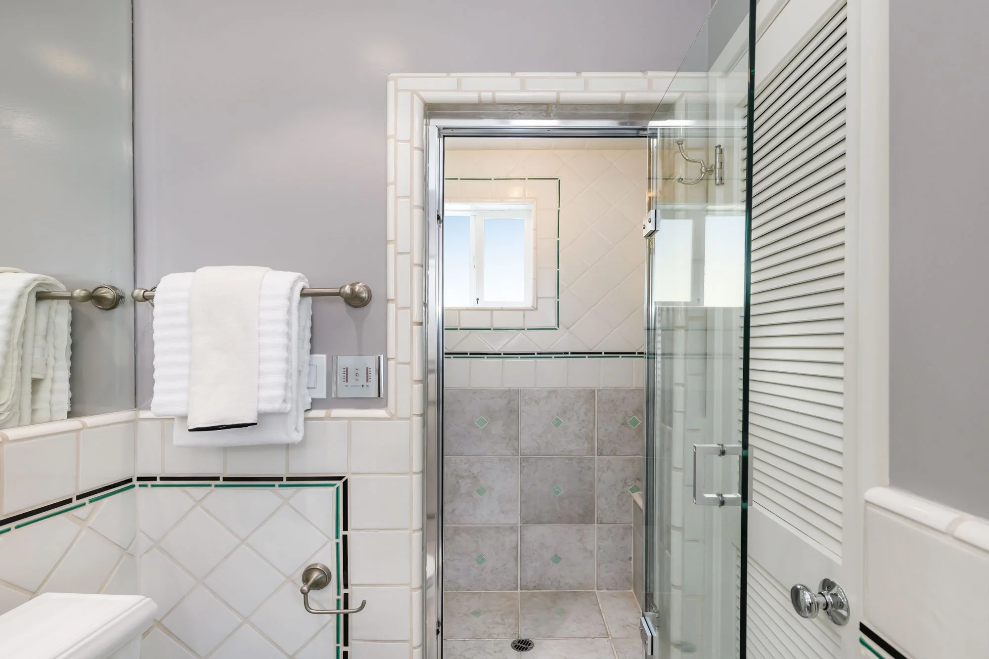 Bathroom with white tiles, towel rack with white towels, small window, glass shower enclosure, and white heating radiator.