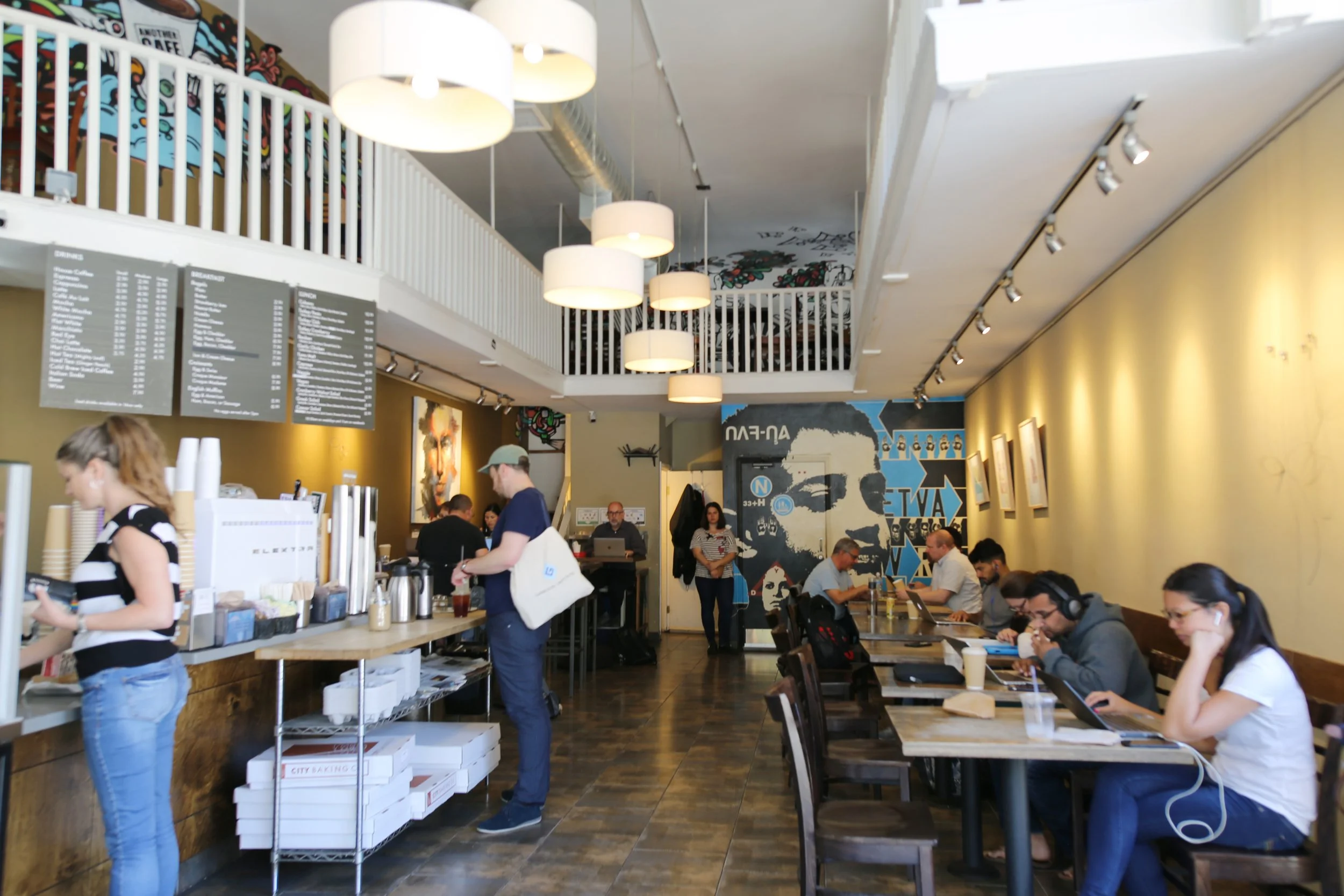 Interior of a busy coffee shop with customers ordering, working on laptops, and socializing. The shop has a double-story design with hanging lights and artwork on the walls.