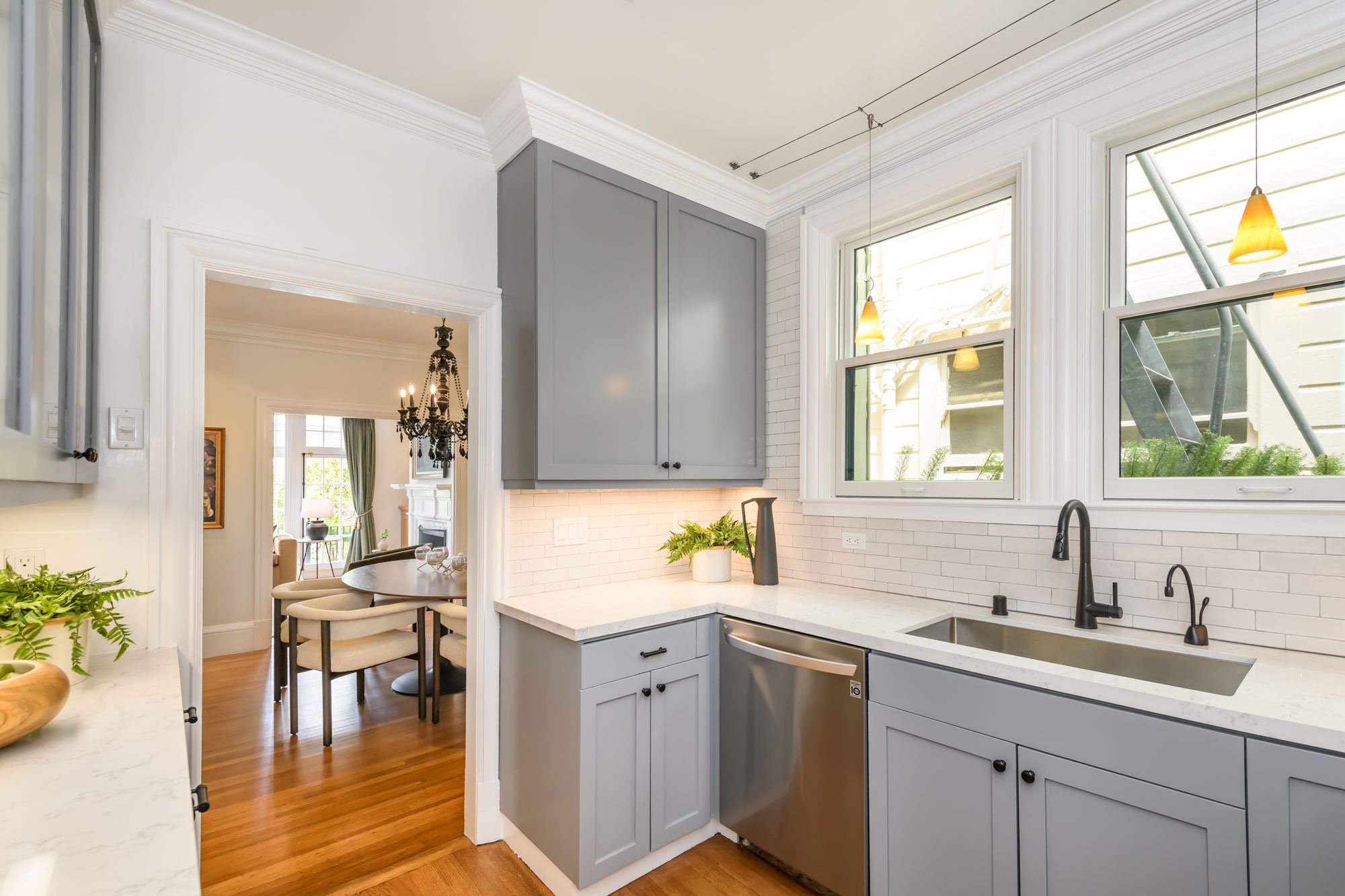 A modern kitchen with white countertops, gray cabinets, and white walls featuring a backsplash of white subway tiles. Large windows let in natural light, and there is a view into a dining area with a chandelier and a round table with chairs. Inside t
