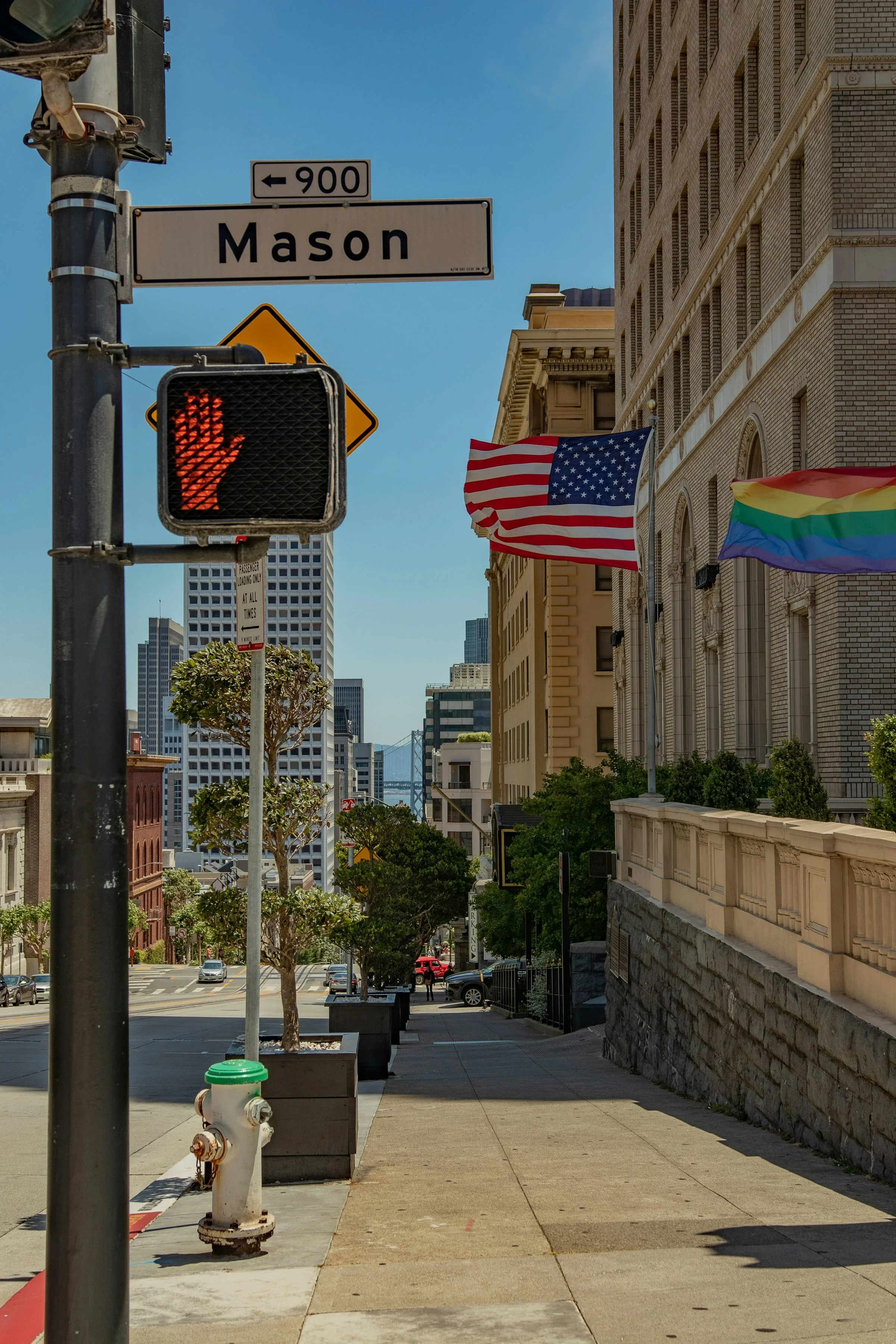 Street view showing a street sign for Mason at an intersection, a pedestrian crosswalk signal with a red hand, American and LGBTQ pride flags hanging from the building, and city buildings in the background on a clear sunny day.