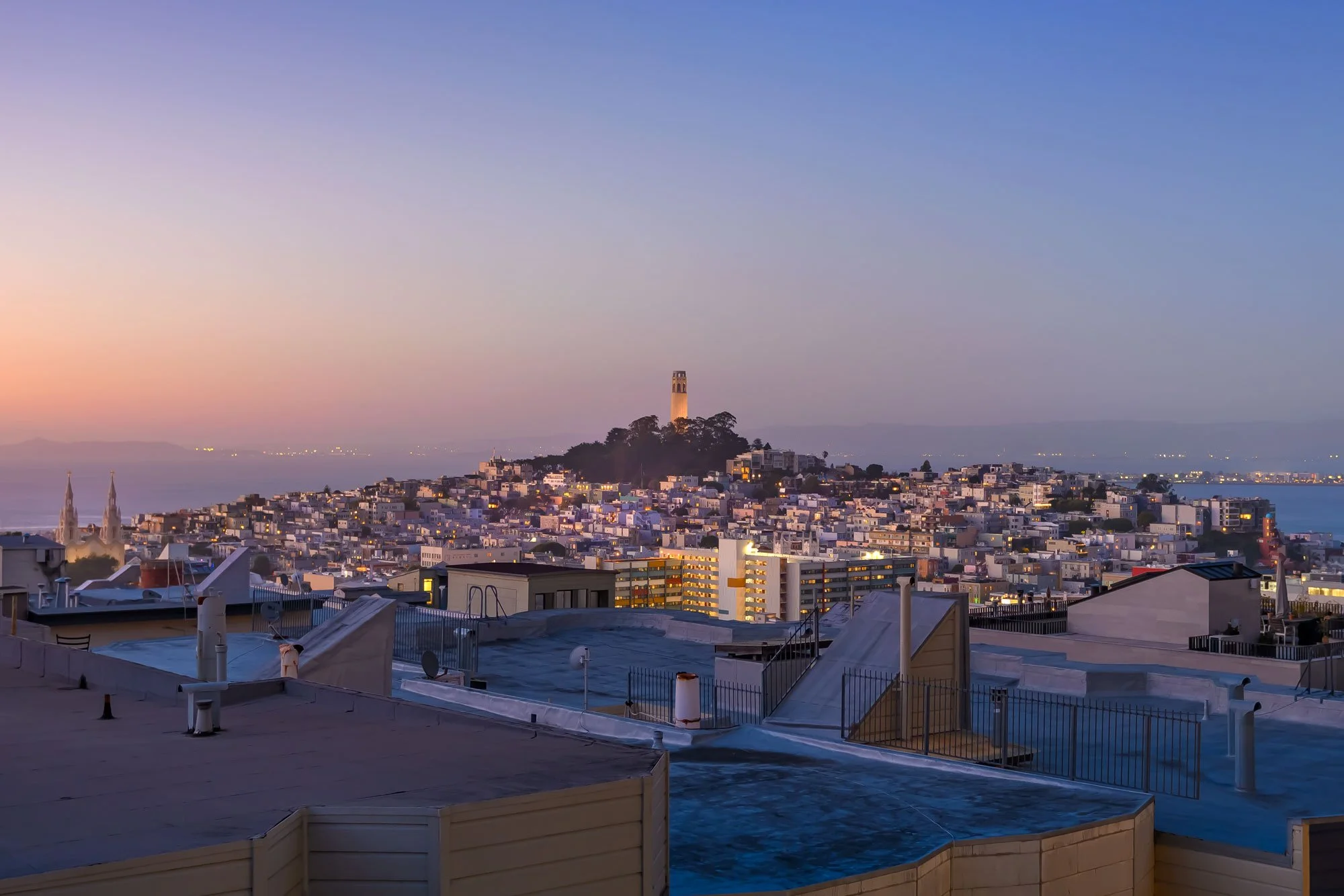 A cityscape at twilight featuring a tall tower on a hill, residential buildings, and a body of water in the background, with pastel-colored sky and city lights starting to turn on.