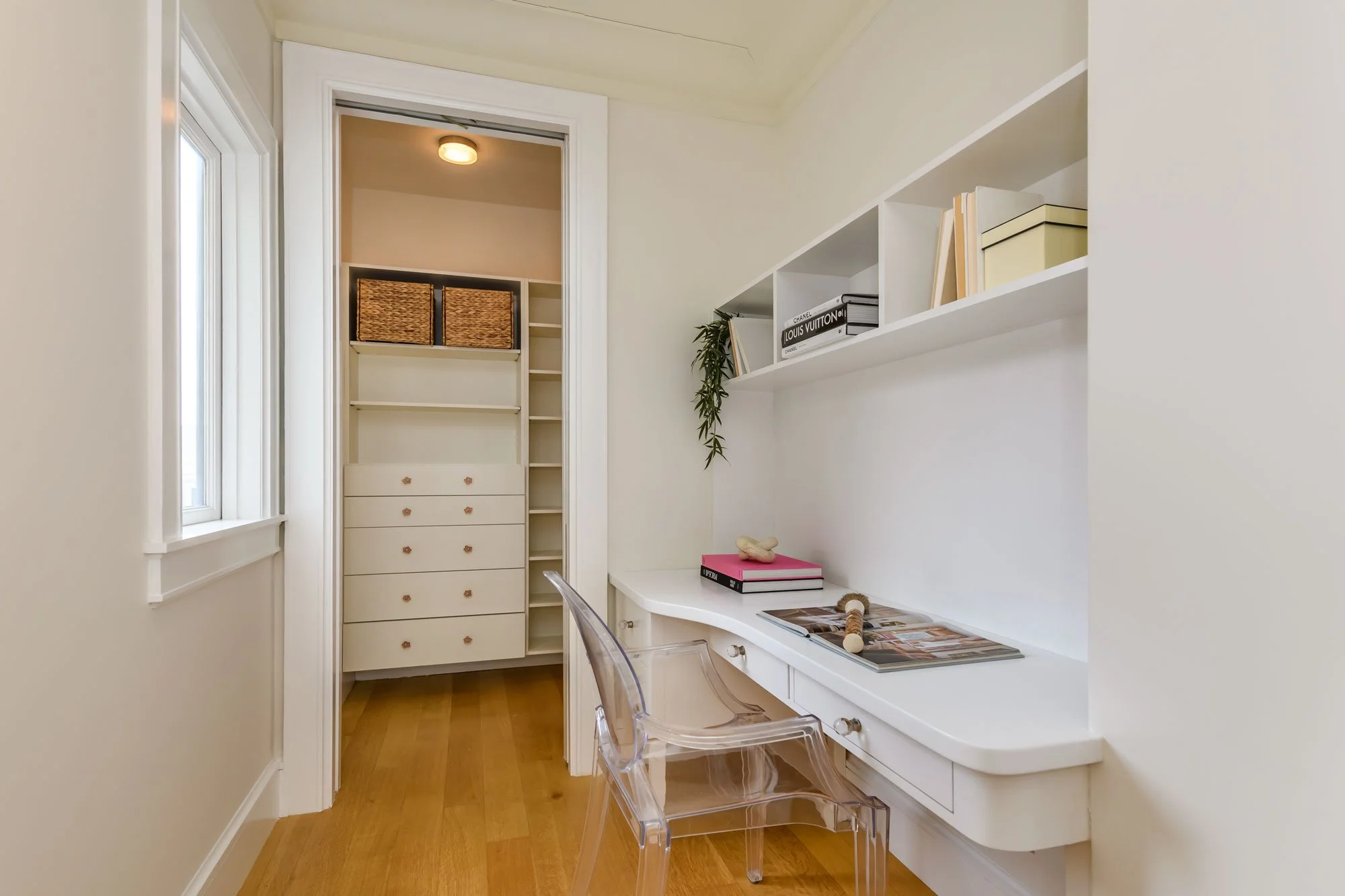 A small home office with a white built-in desk, acrylic chair, and white shelving with books, boxes, and plants, next to a window with blinds.