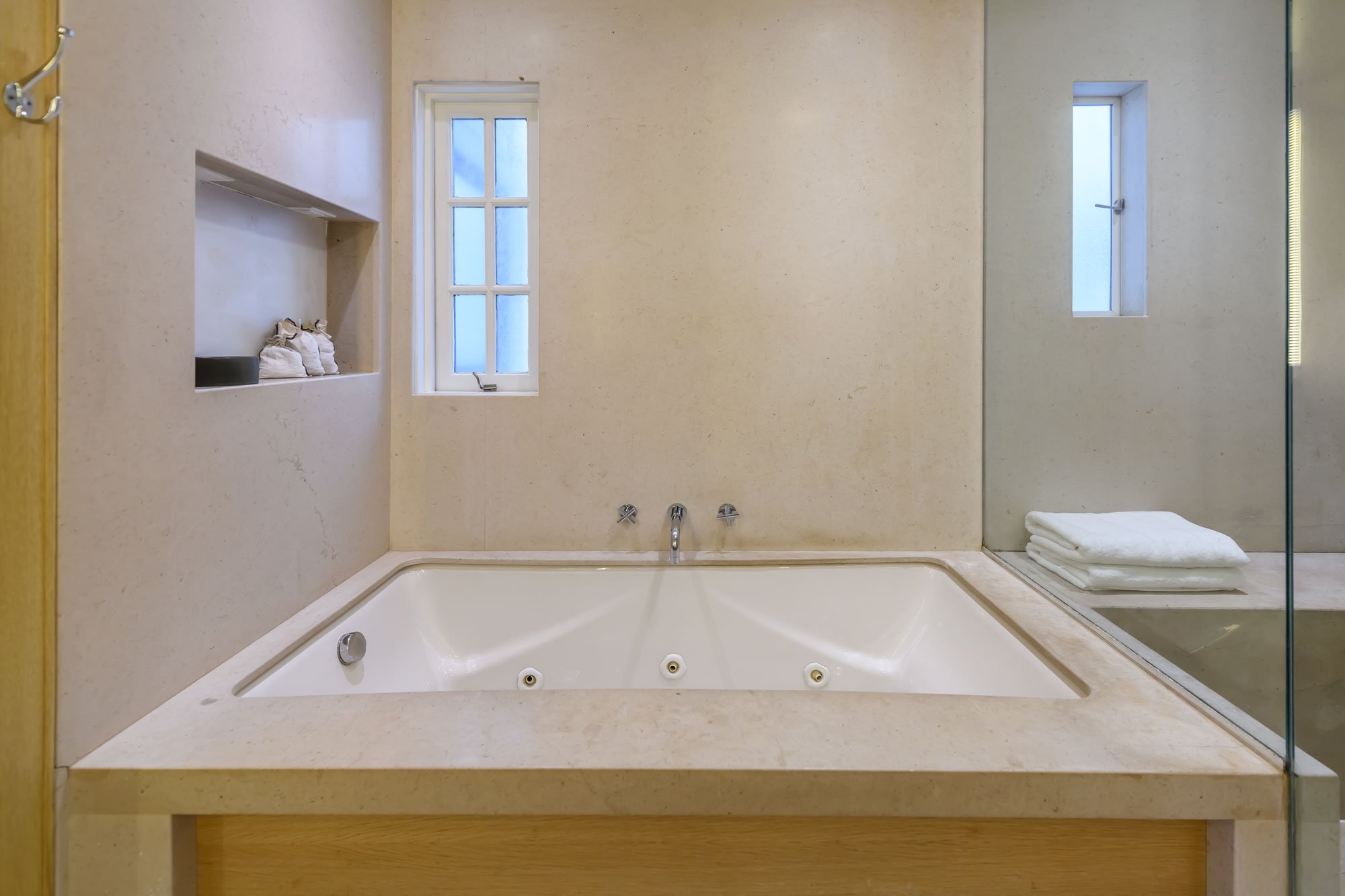 Bathroom with a built-in bathtub, two small windows, a shelf with toiletries, and a folded towel on a shelf behind a glass shower enclosure.