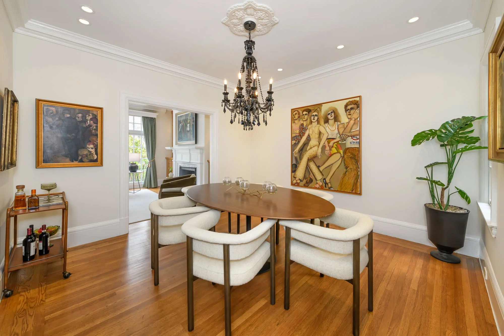 A dining room with a wooden table surrounded by beige upholstered chairs. The room features a chandelier, artwork on the walls, and a potted monstera plant. A doorway opens to a living room with a fireplace and large window.