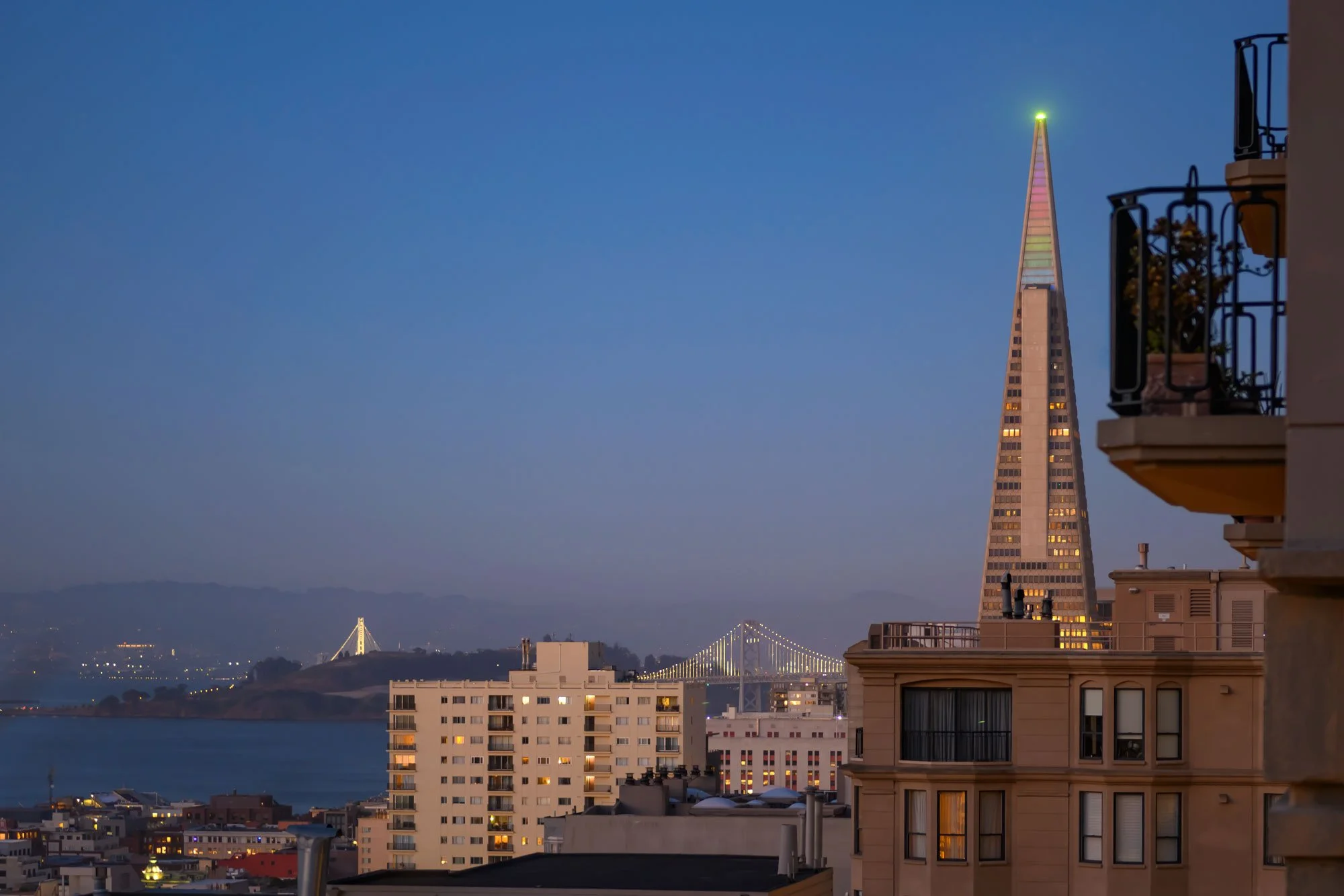 Cityscape at dusk featuring the illuminated Salesforce Tower with a colorful spire, Bay Bridge with lights, and waterfront buildings in San Francisco.