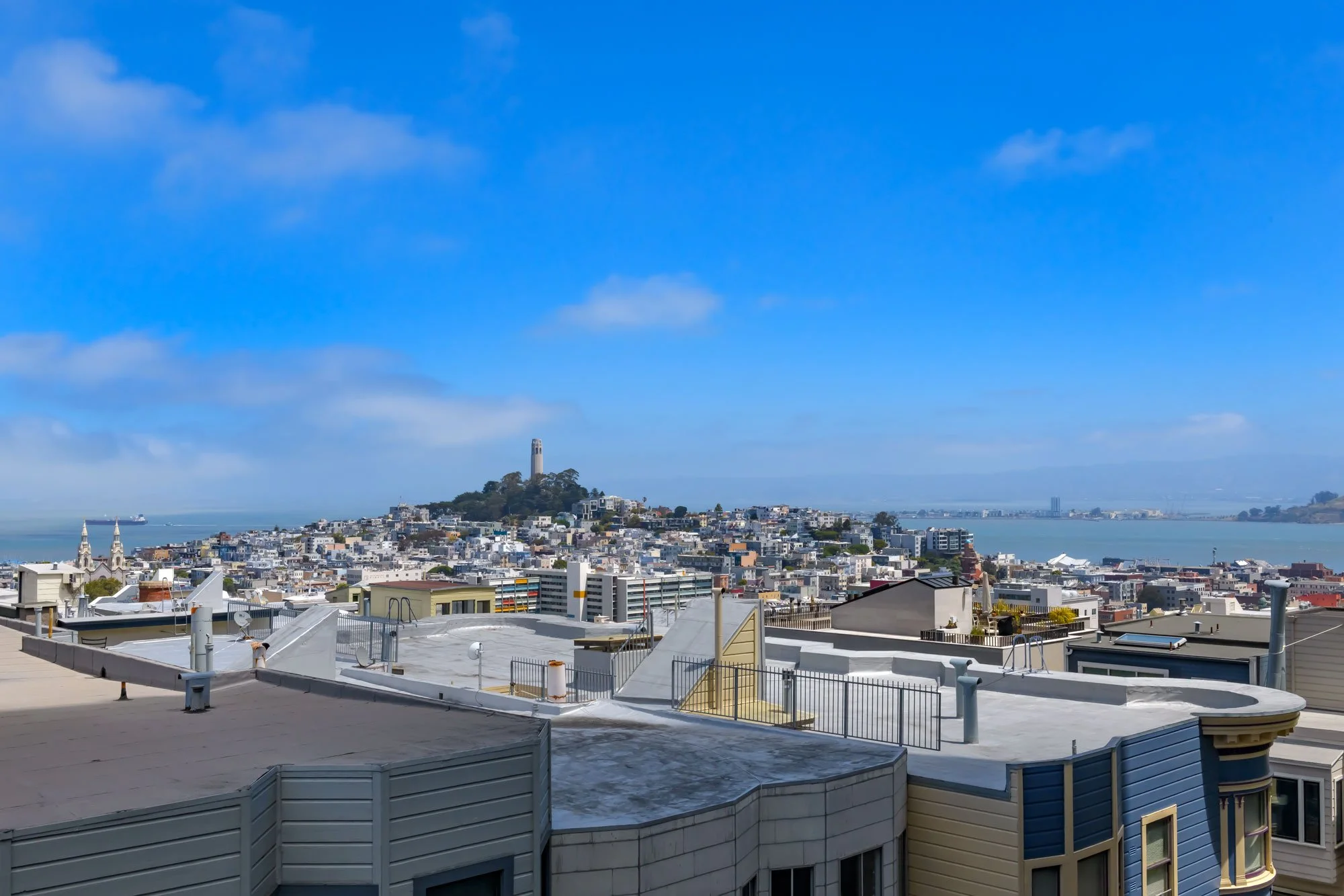 Cityscape with buildings, a hill with a tower, water in the background, and a clear blue sky with some clouds.